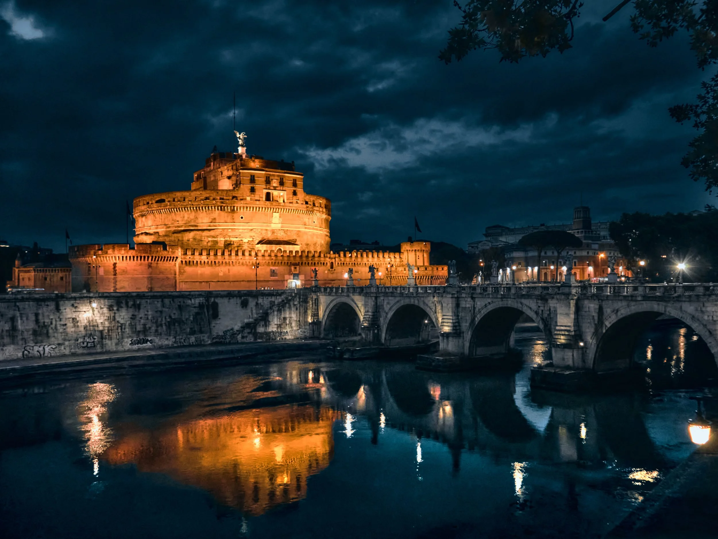 Castel Sant'Angelo iluminado por la noche reflejado en el río Tíber en Roma, con cielo nublado y luces en el fondo.