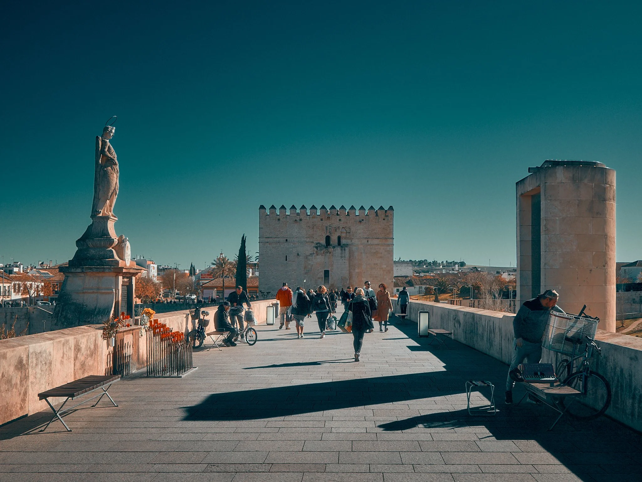 Puente con estatuas y personas caminando, con edificio antiguo en fondo y cielo despejado.