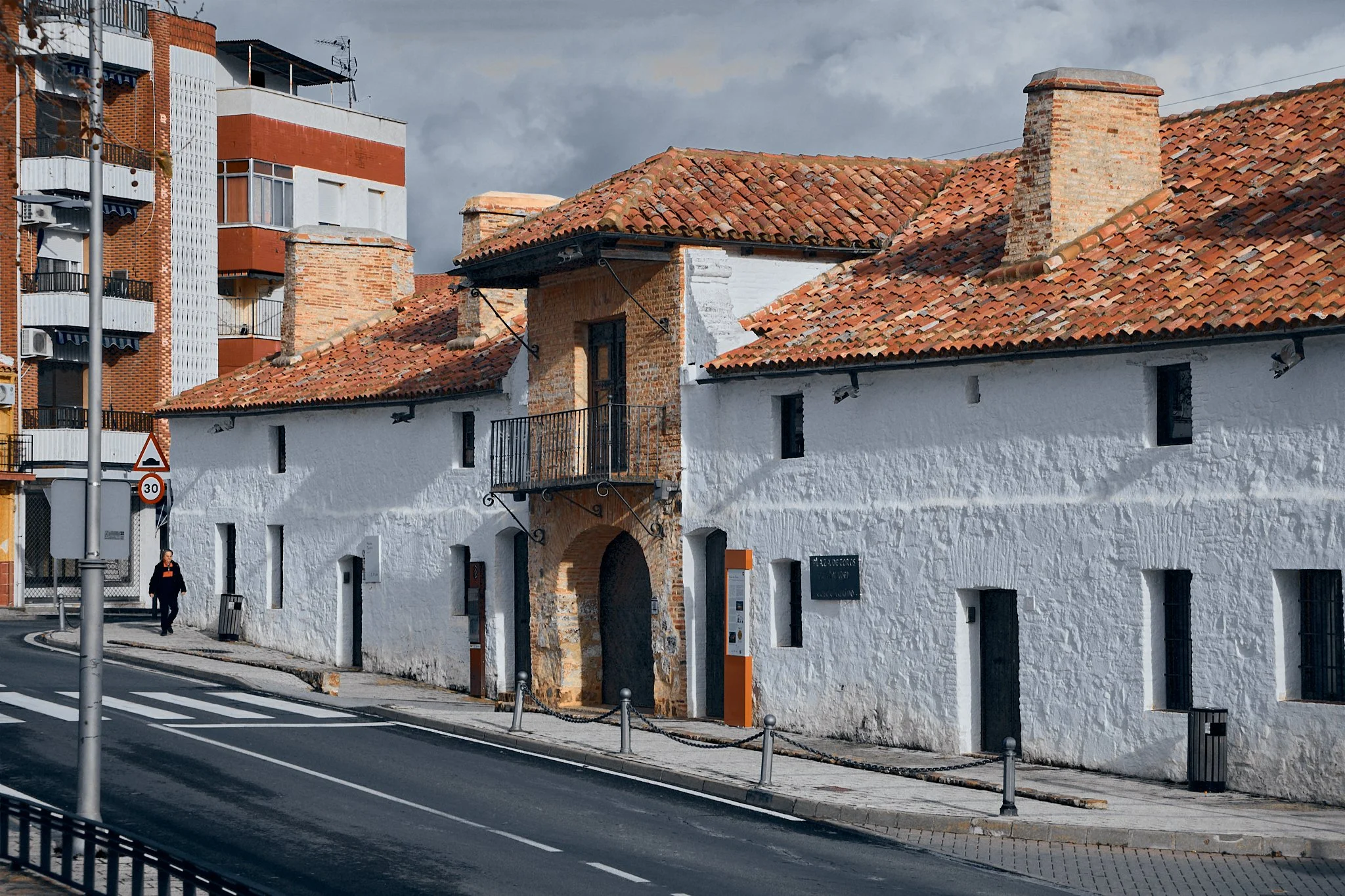 Casa antigua con paredes blancas y techos de tejas rojas, ubicada en una calle urbana, con un arco en la entrada y una baca en la parte superior. Un área moderna de edificios altos se observa al fondo.