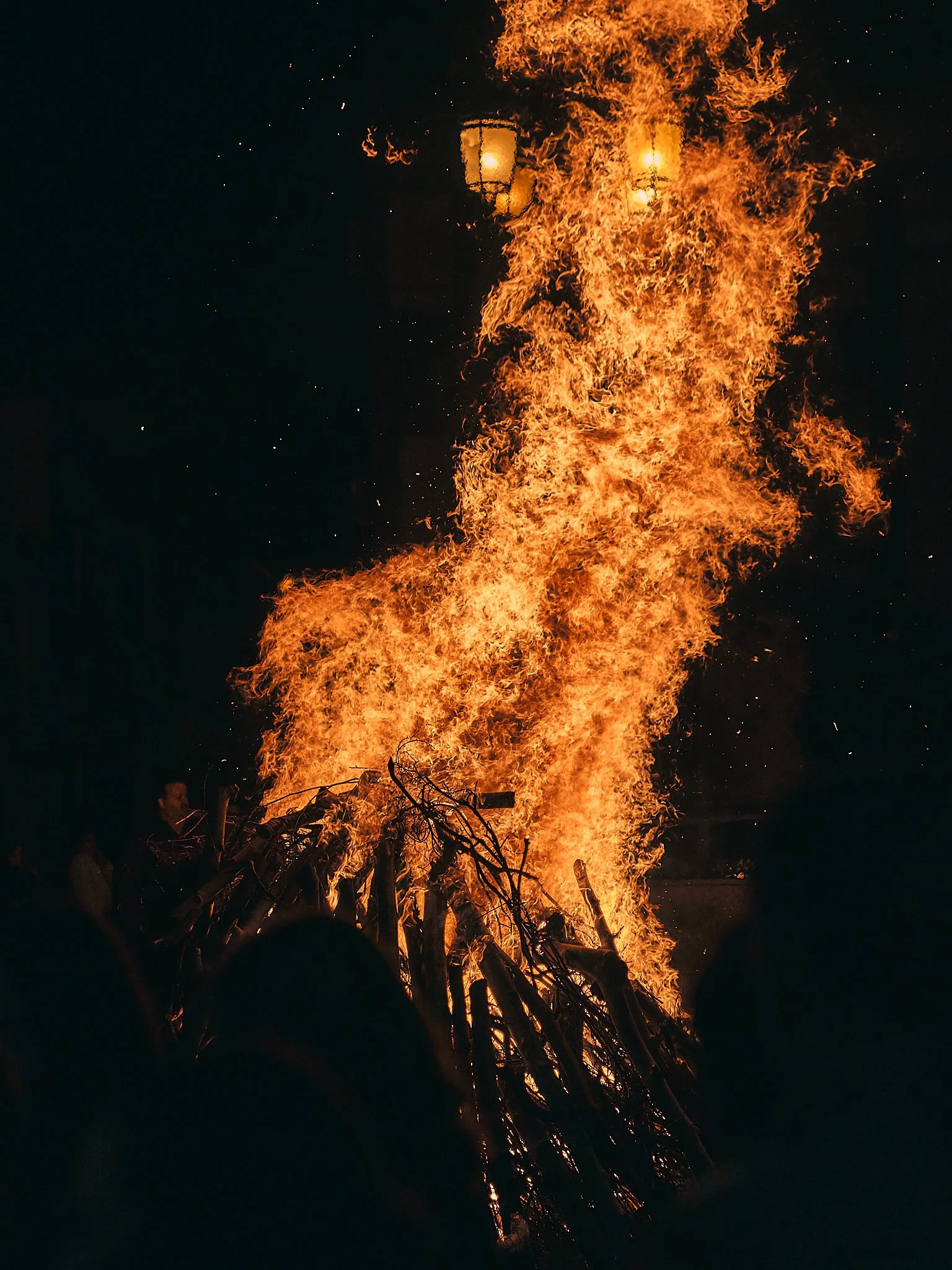 Una hoguera grande de madera y ramas ardiendo durante la noche, con llamas altas y chispas que salen del fuego.