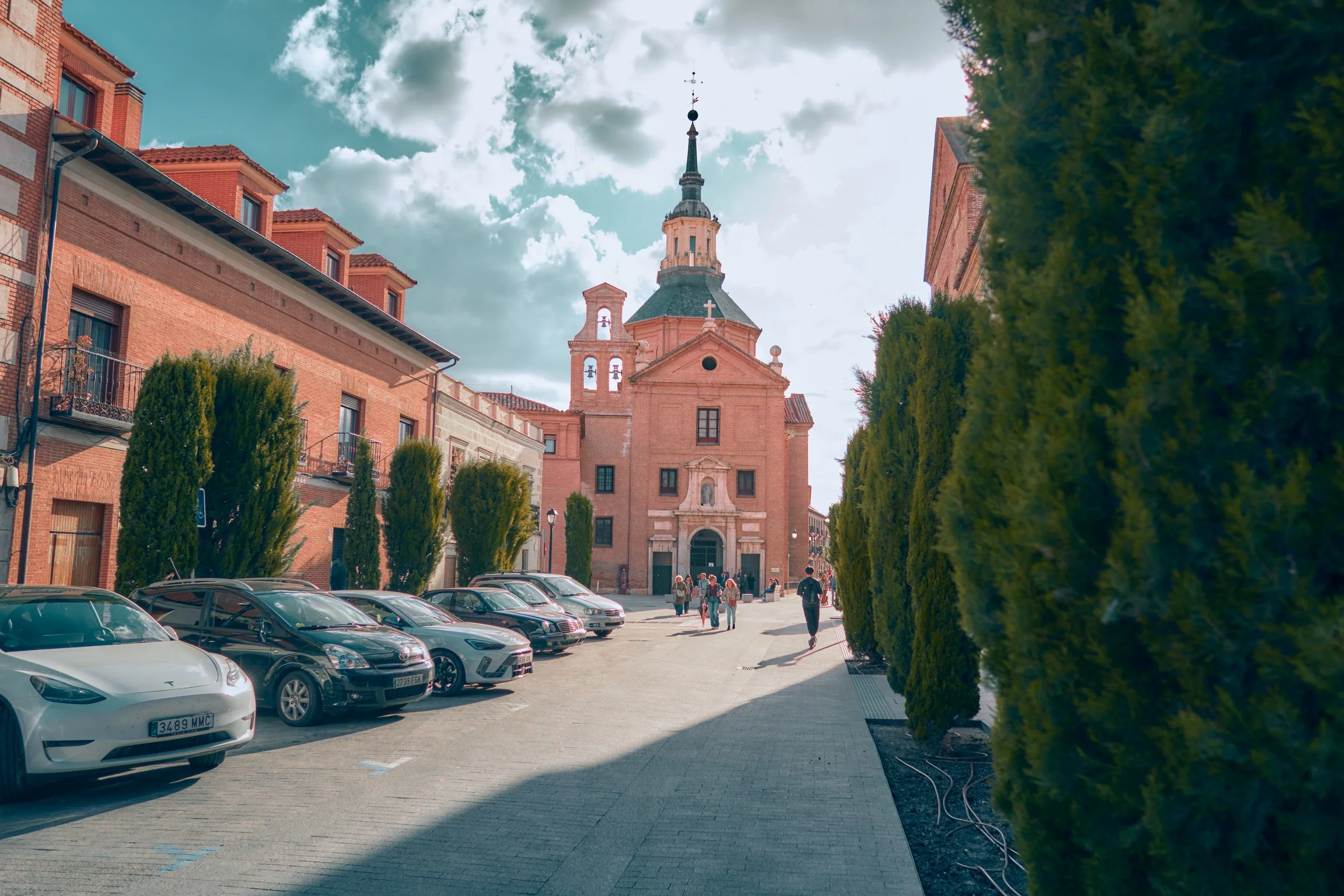 Calle con árboles a ambos lados, coches estacionados y una iglesia de ladrillo al fondo