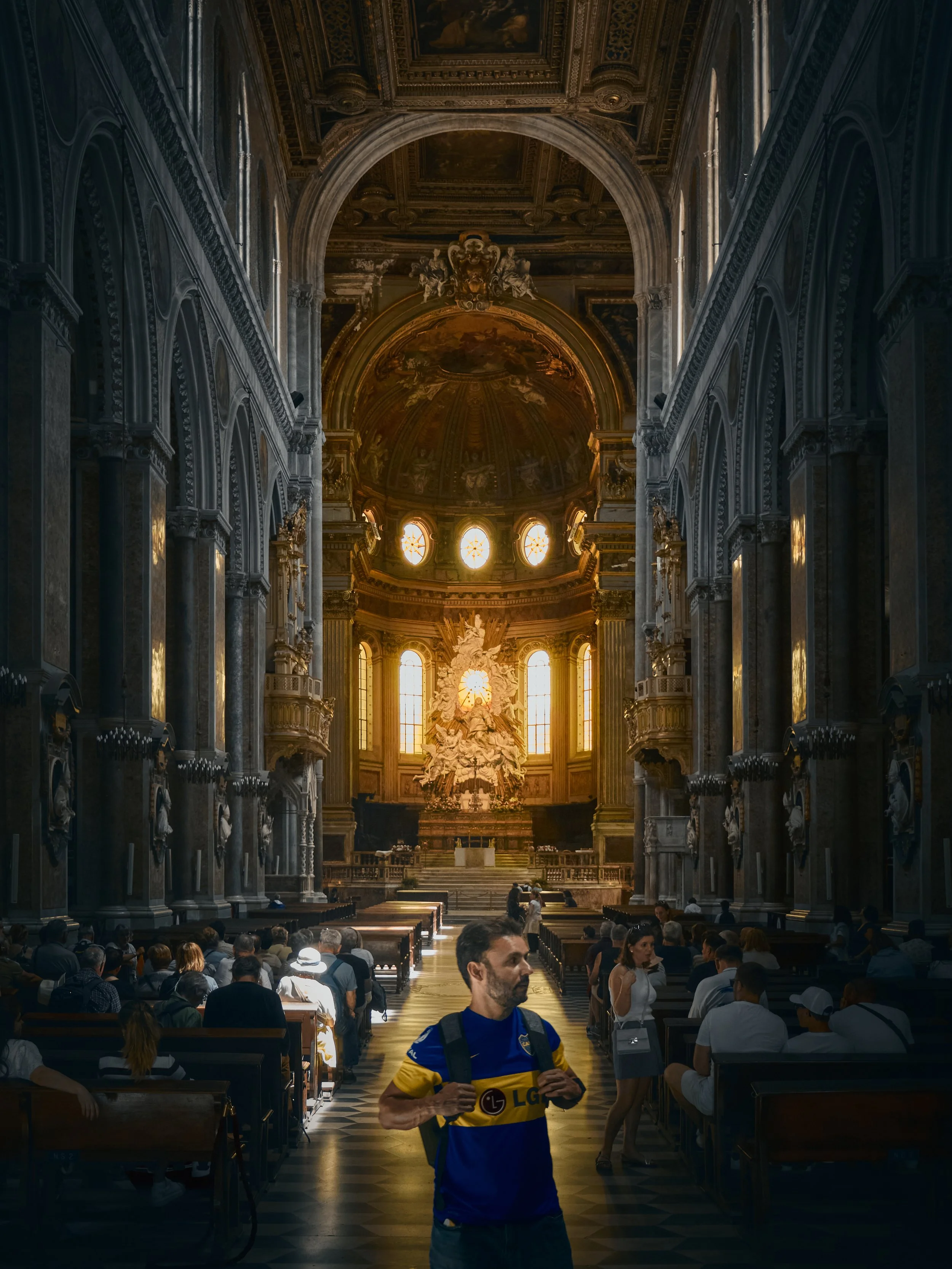 Interior de una iglesia o catedral con muchos asistentes, un hombre con camiseta azul y amarillo con mochila en primer plano, y en el altar una escultura blanca y dorada con ventanas iluminadas al fondo.