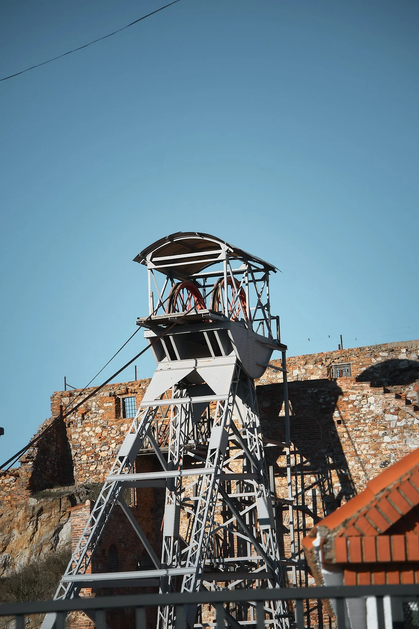 Estructura metálica de un antiguo telégrafo o sistema de transmisión, con ruedas en la parte superior, en un entorno rocoso con restos de una muralla de ladrillos y una casa con techo de tejas al fondo, bajo un cielo despejado.