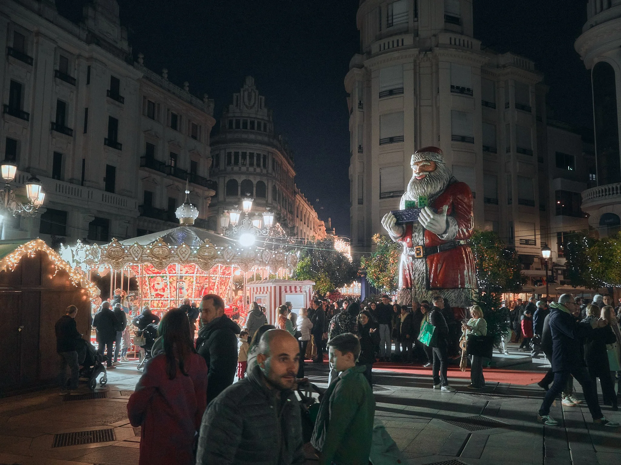 Escena nocturna en una plaza con decoraciones navideñas, incluyendo un gran muñeco de Santa Claus con un cuaderno y una caja, y un carrusel iluminado en el fondo, con varias personas caminando y disfrutando del ambiente festivo.