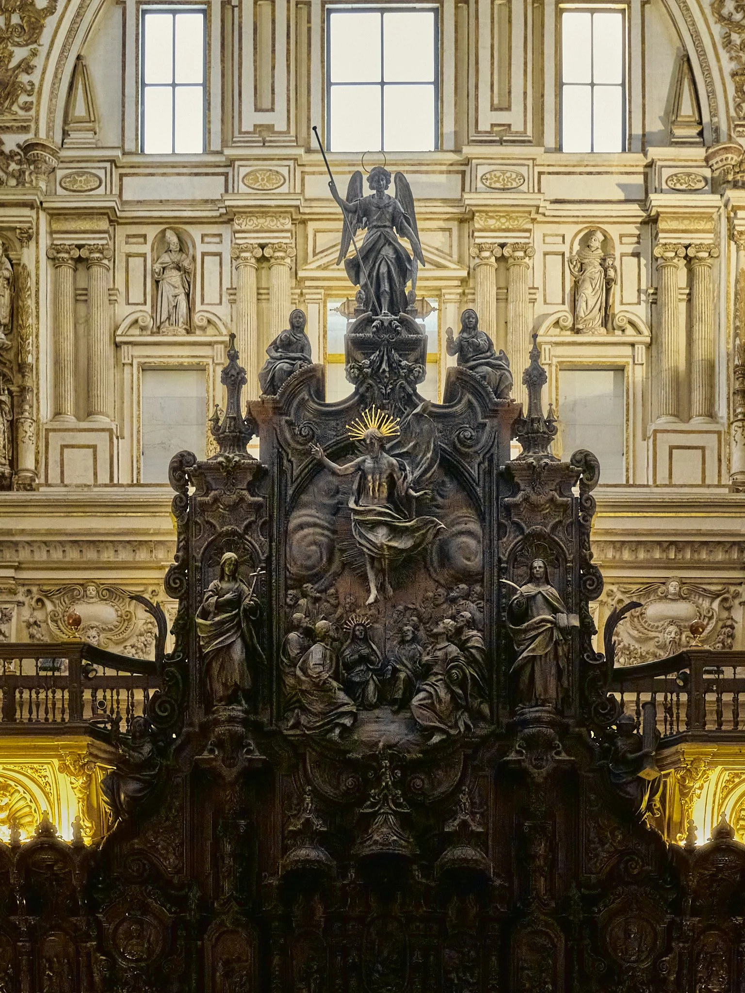 Altarpiece en una iglesia con esculturas religiosas en tonos dorados y oscuros, incluyendo figuras de santos y Dios, bajo ventanas grandes.