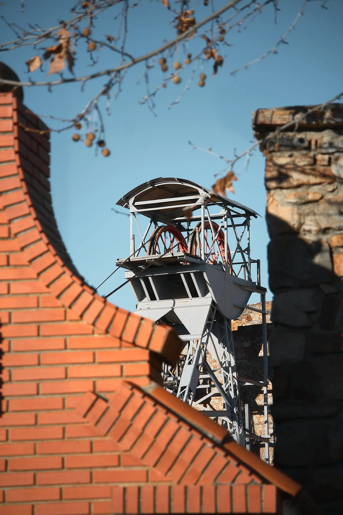 Una torre de vigilancia antigua vista entre techos de tejas y muros de piedra, con ramas de árbol sin hojas en primer plano y un cielo azul de fondo.