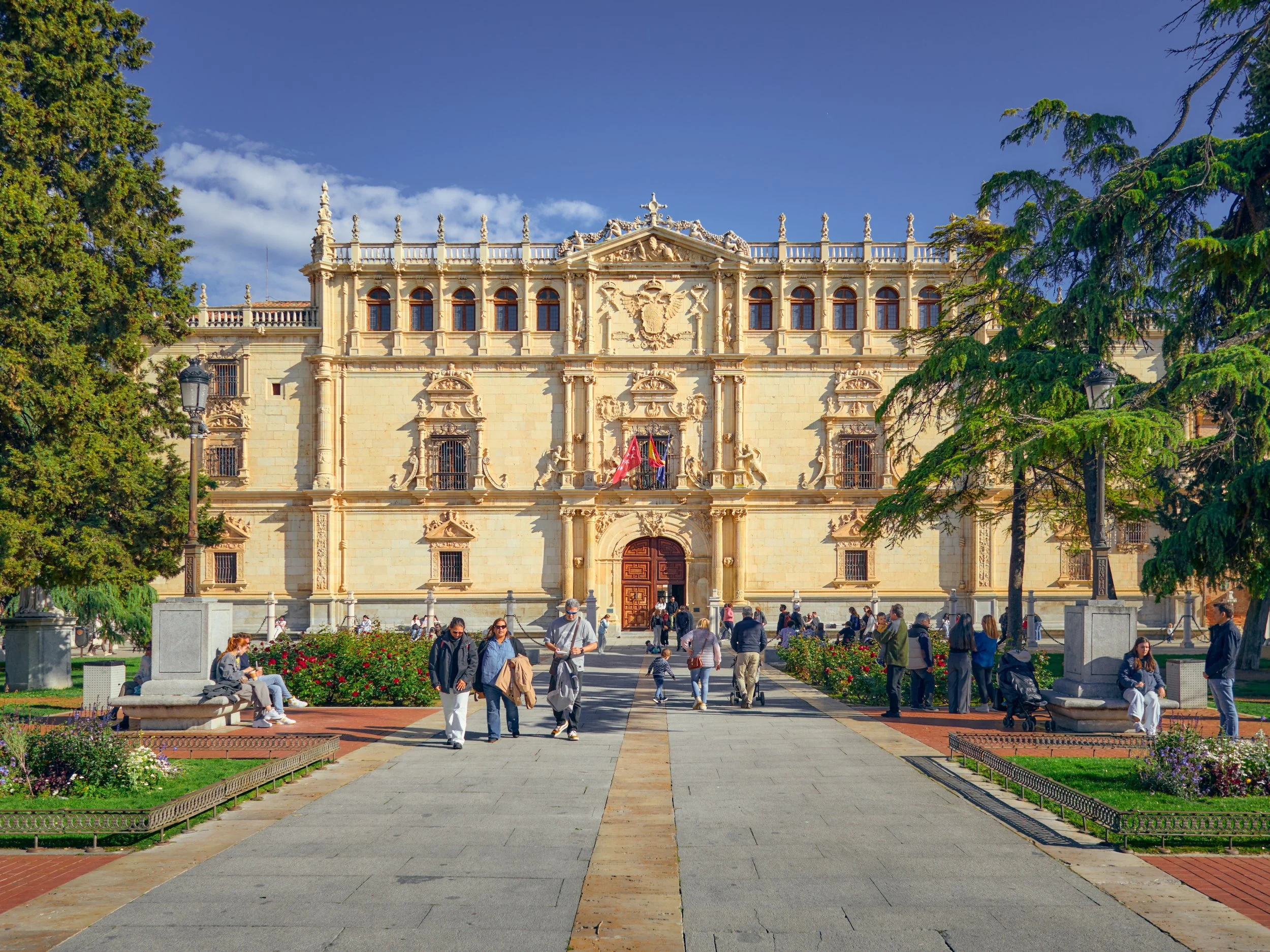 Edificio histórico de color beige con detalles arquitectónicos elaborados, rodeado de áreas verdes y personas caminando y sentadas en el parque frente a él.