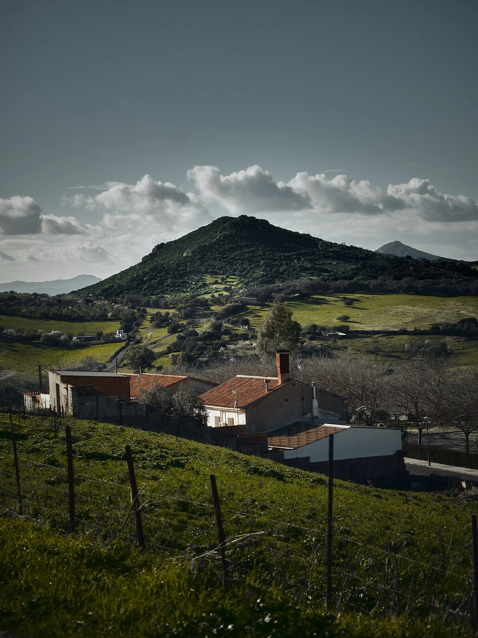 Vista de un paisaje rural con casas blancas y techos de teja, colinas verdes y una montaña en el fondo, cielo parcialmente nublado.