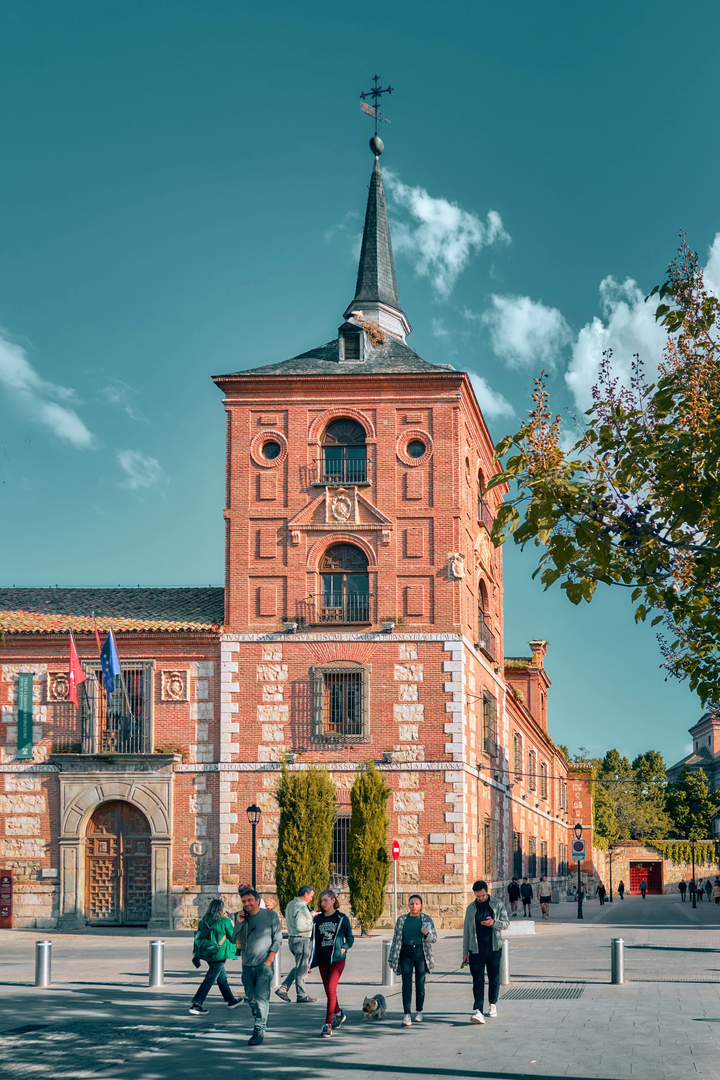 Una iglesia de ladrillo con una torre y un campanario, varias personas caminan frente a ella en una calle, árboles y cielo con nubes