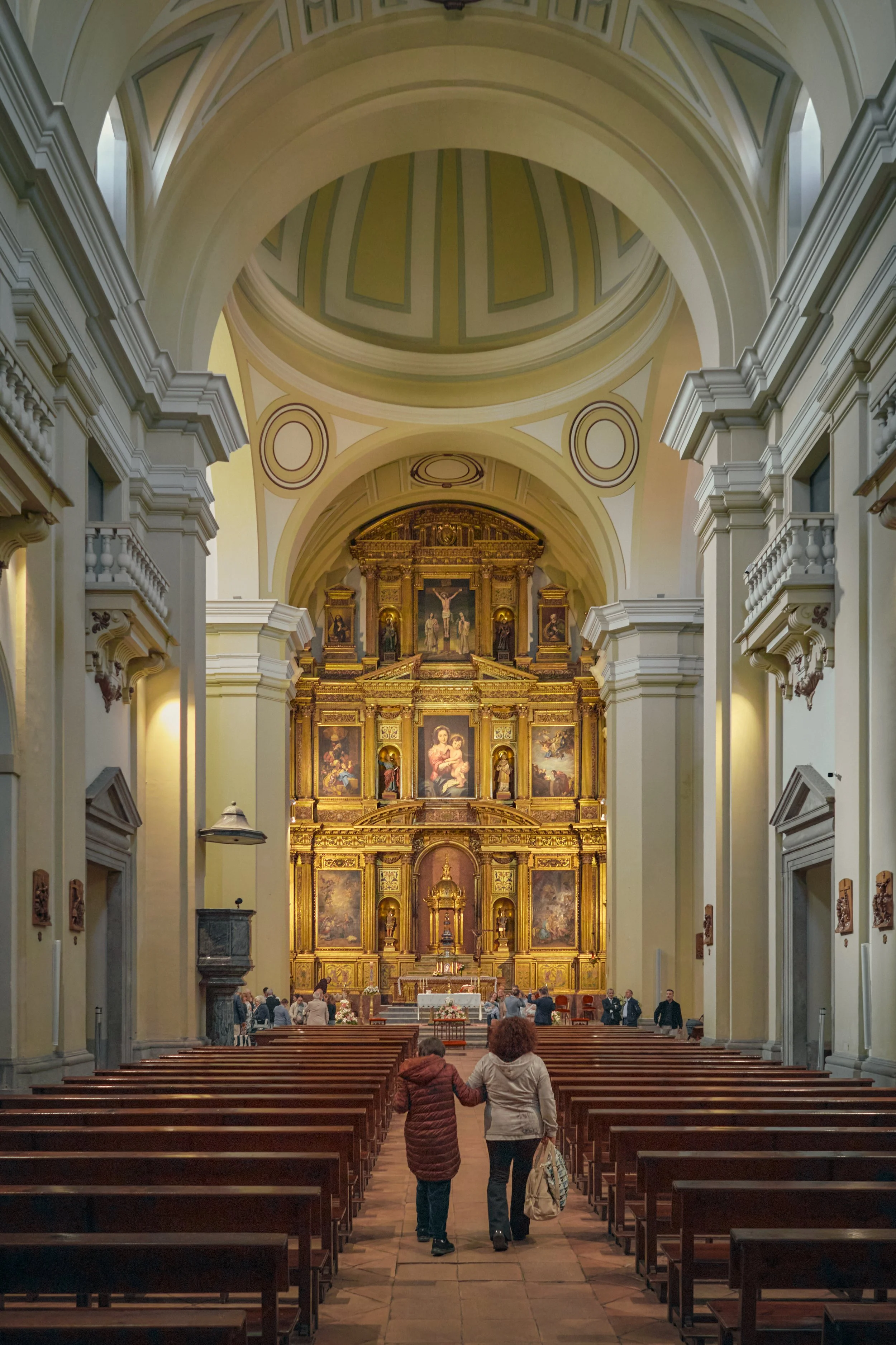 Interior de una iglesia con altos arcos y altar dorado con pinturas religiosas, dos personas caminan entre los bancos de madera.