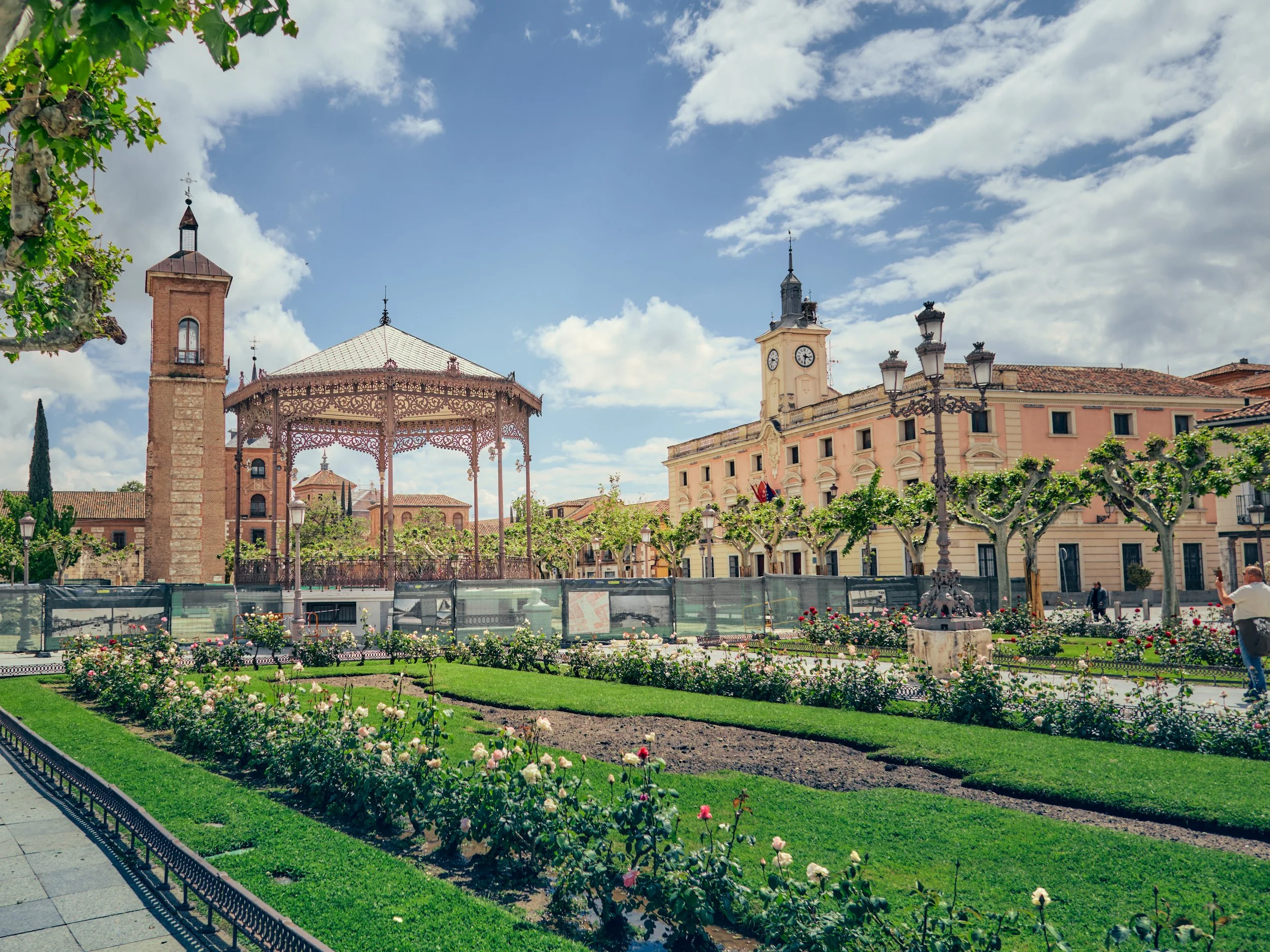 Plaza con jardines, fuentes, y estructuras históricas, con edificios y un cielo parcialmente nublado en el fondo.