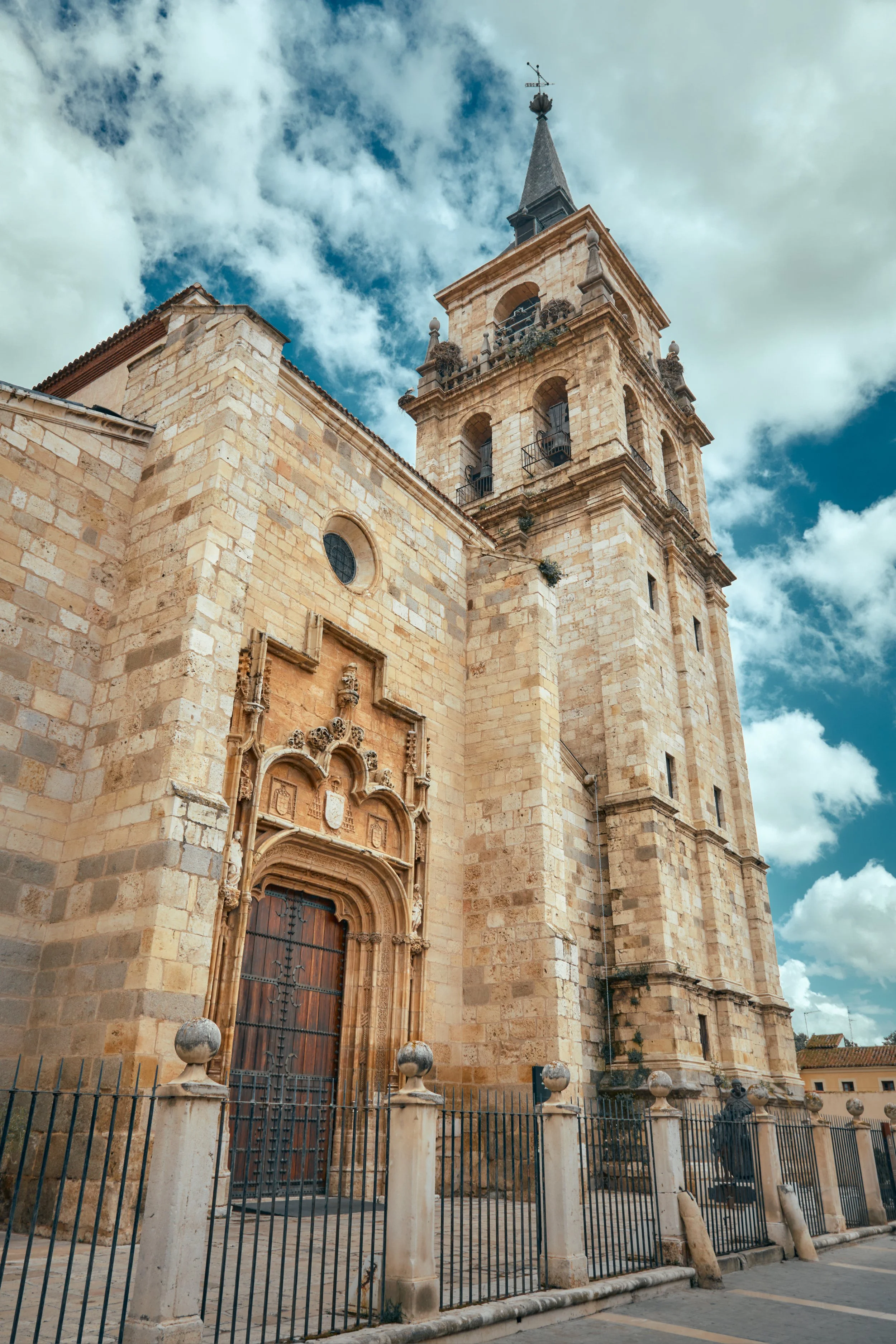 Vista frontal de una iglesia de piedra con torre y campanario, puertas de madera tallada, reja negra y cielo con nubes.