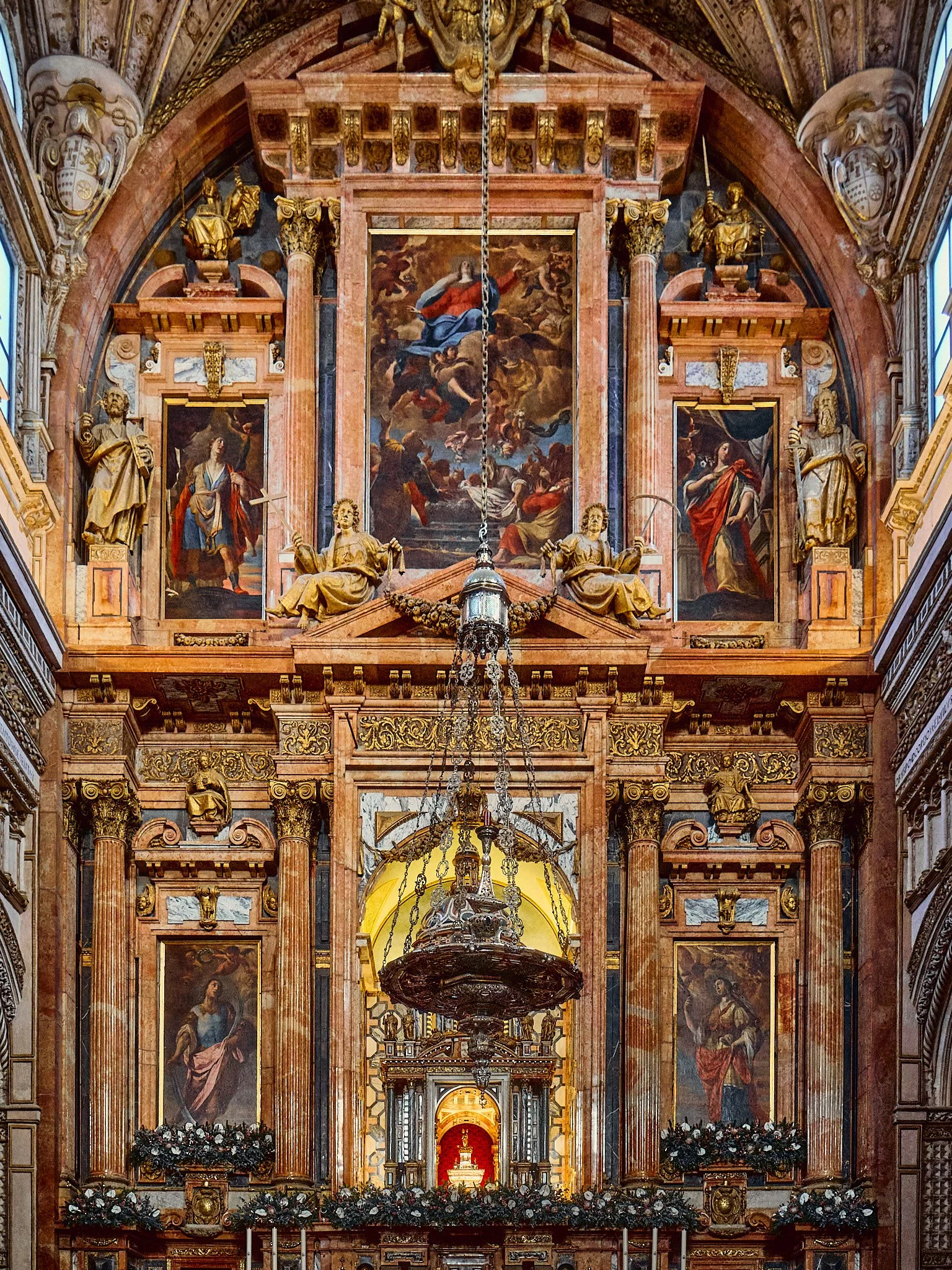 Interior de una iglesia barroca con pinturas y esculturas doradas, columnas de mármol y un candelabro suspendido en el centro.