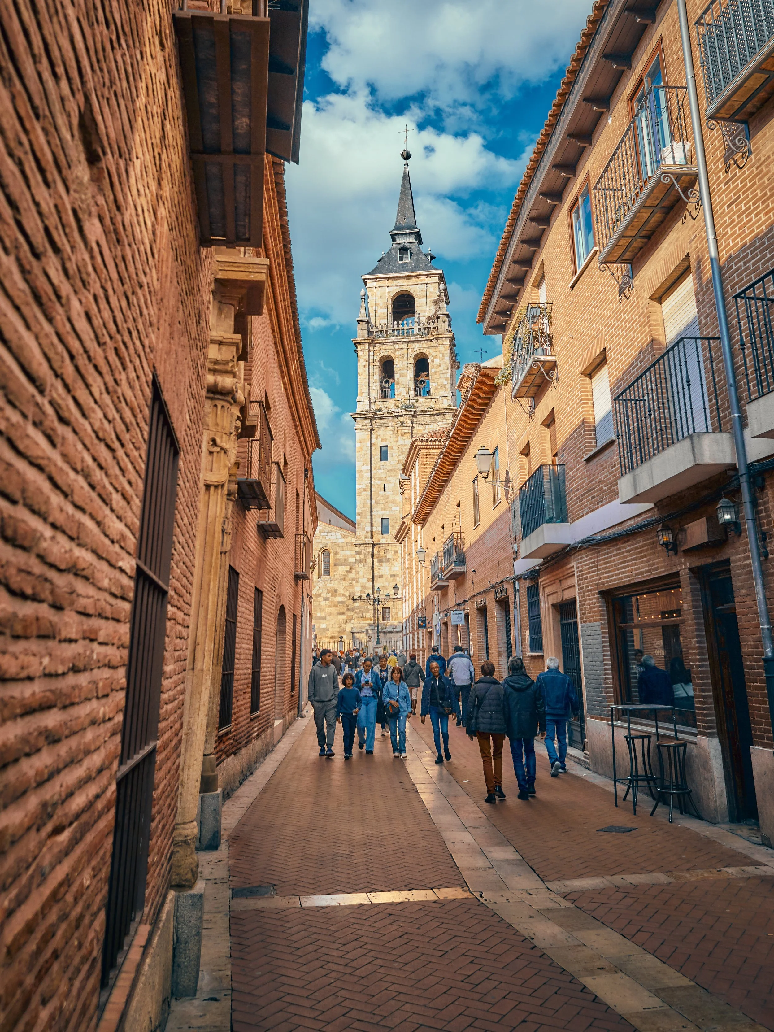 Calle estrecha con ladrillos y edificios de estilo colonial, una torre de iglesia con reloj y una cruz en la cima, cielo azul con nubes y gente caminando.