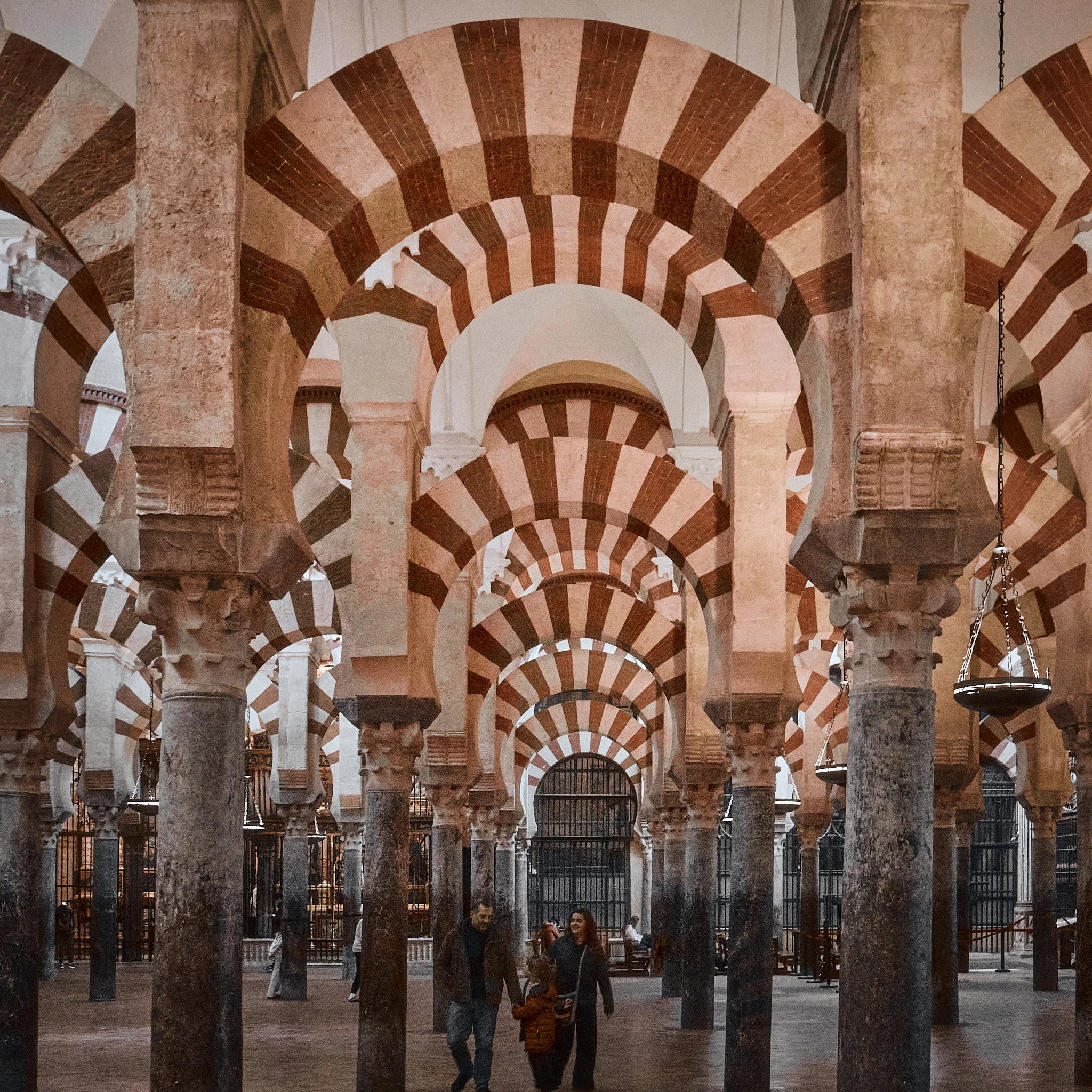 Interior de una iglesia con arcos de piedra y columnas con capiteles decorativos, varias personas caminando, barredales de hierro y bancos.