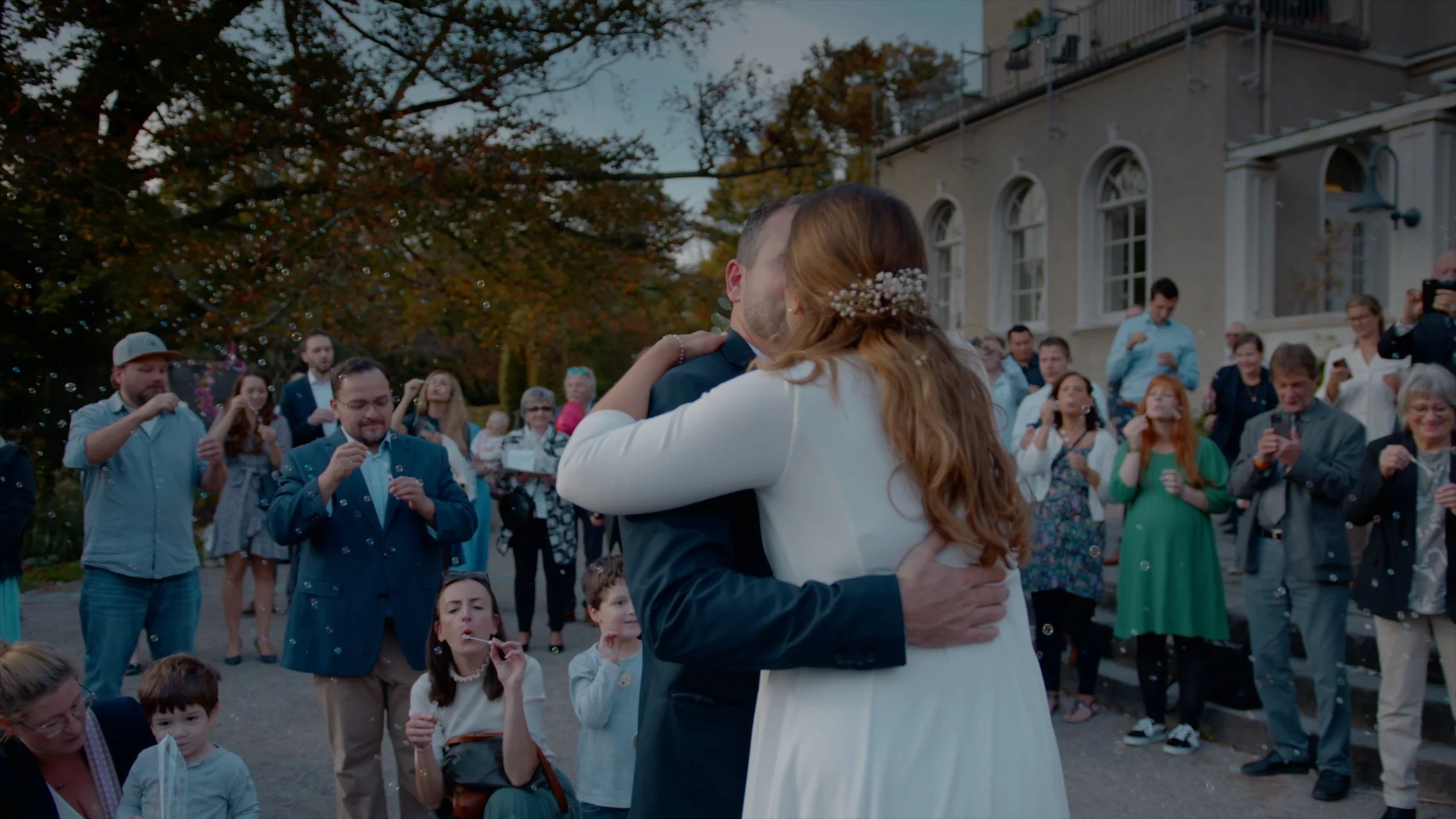 Ein Brautpaar umarmt sich bei einer Hochzeit im Freien, umgeben von Gästen, die Fotos machen und Seifenblasen blasen, in einem historischen Gebäude im Hintergrund, bei Sonnenuntergang.