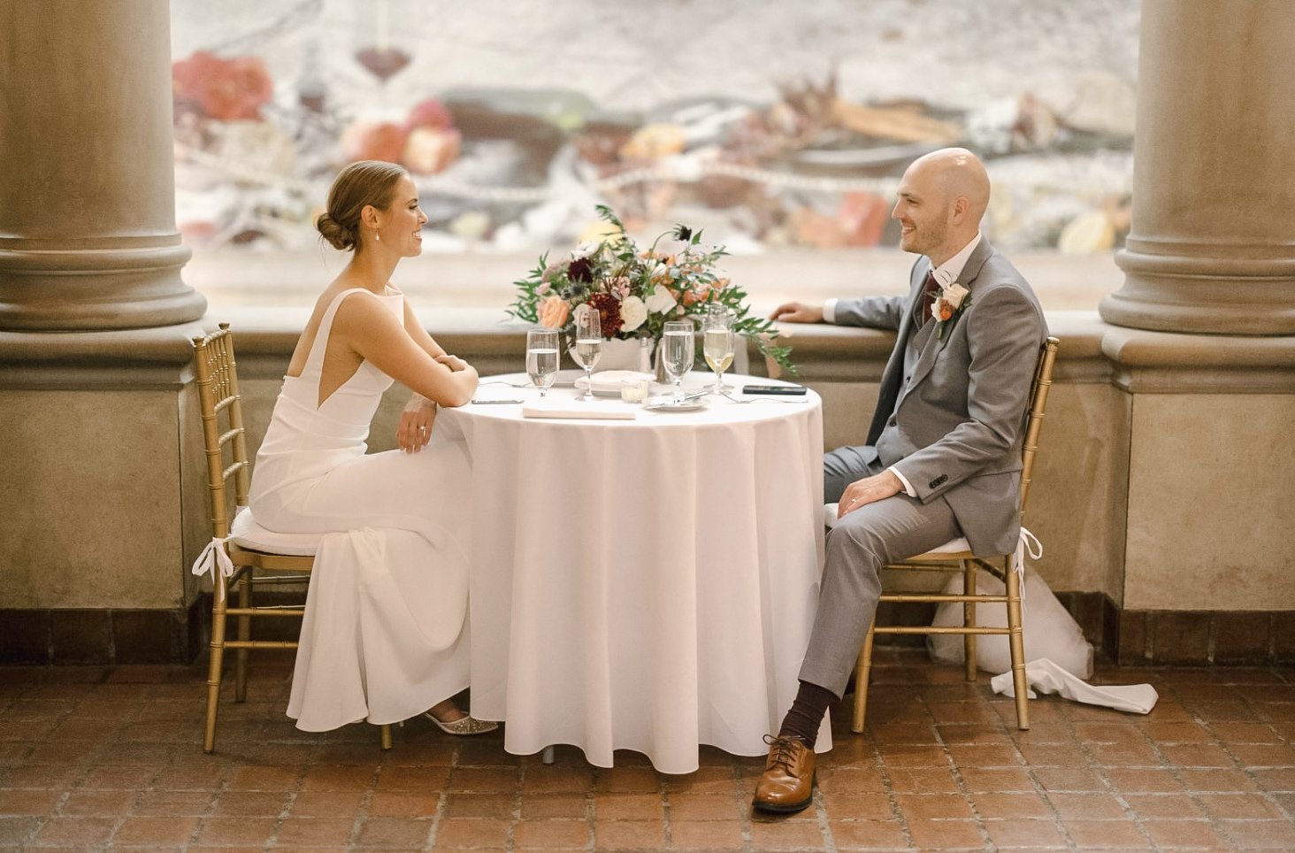 Bride and groom seated at a white-clothed table, sharing an intimate moment with elegant floral centerpiece, formal attire, and elegant indoor setting.