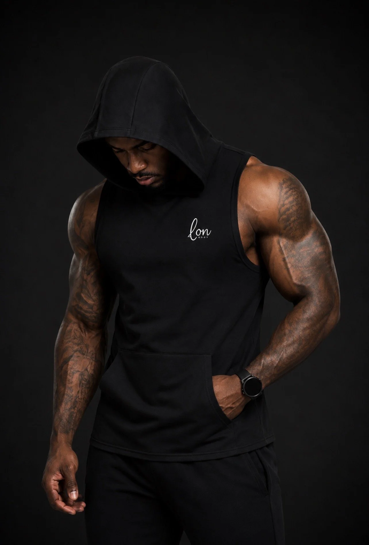 A muscular man wearing a black sleeveless hoodie with the word 'lon' on it, looking down with his left hand in his pocket, against a black background.