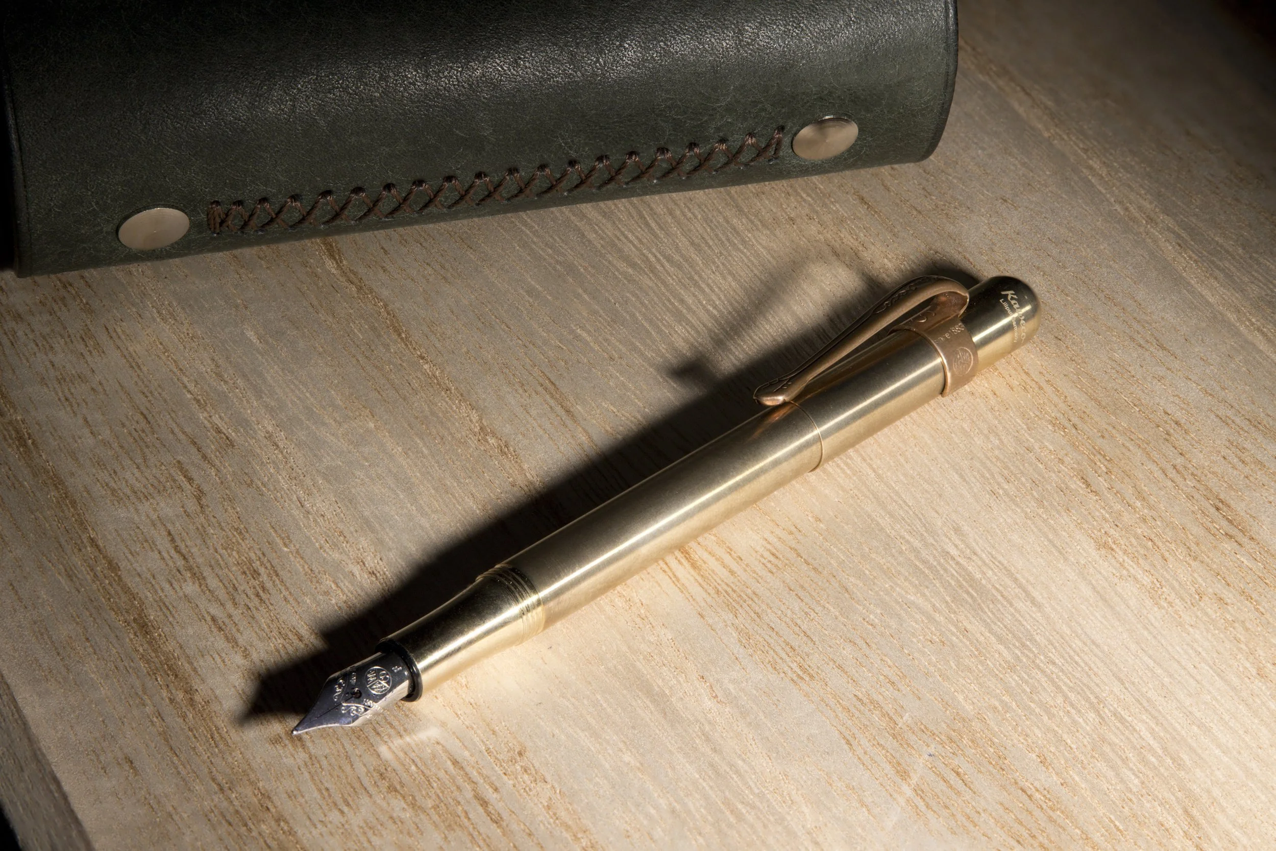 A gold-colored fountain pen resting on a wooden surface near a black leather bag.