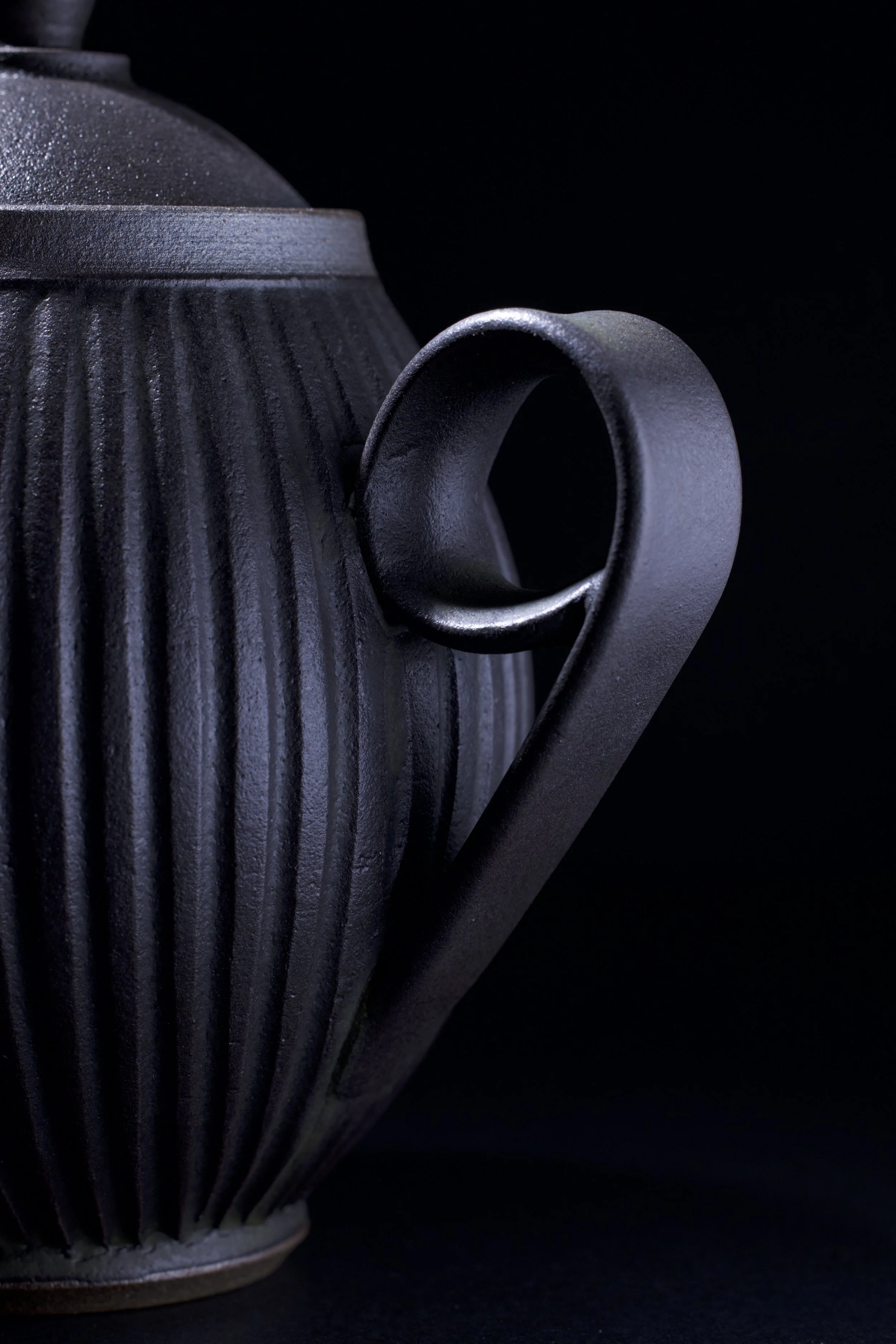 Close-up of a dark gray matte ceramic teapot with a textured ribbed surface, set against a black background.