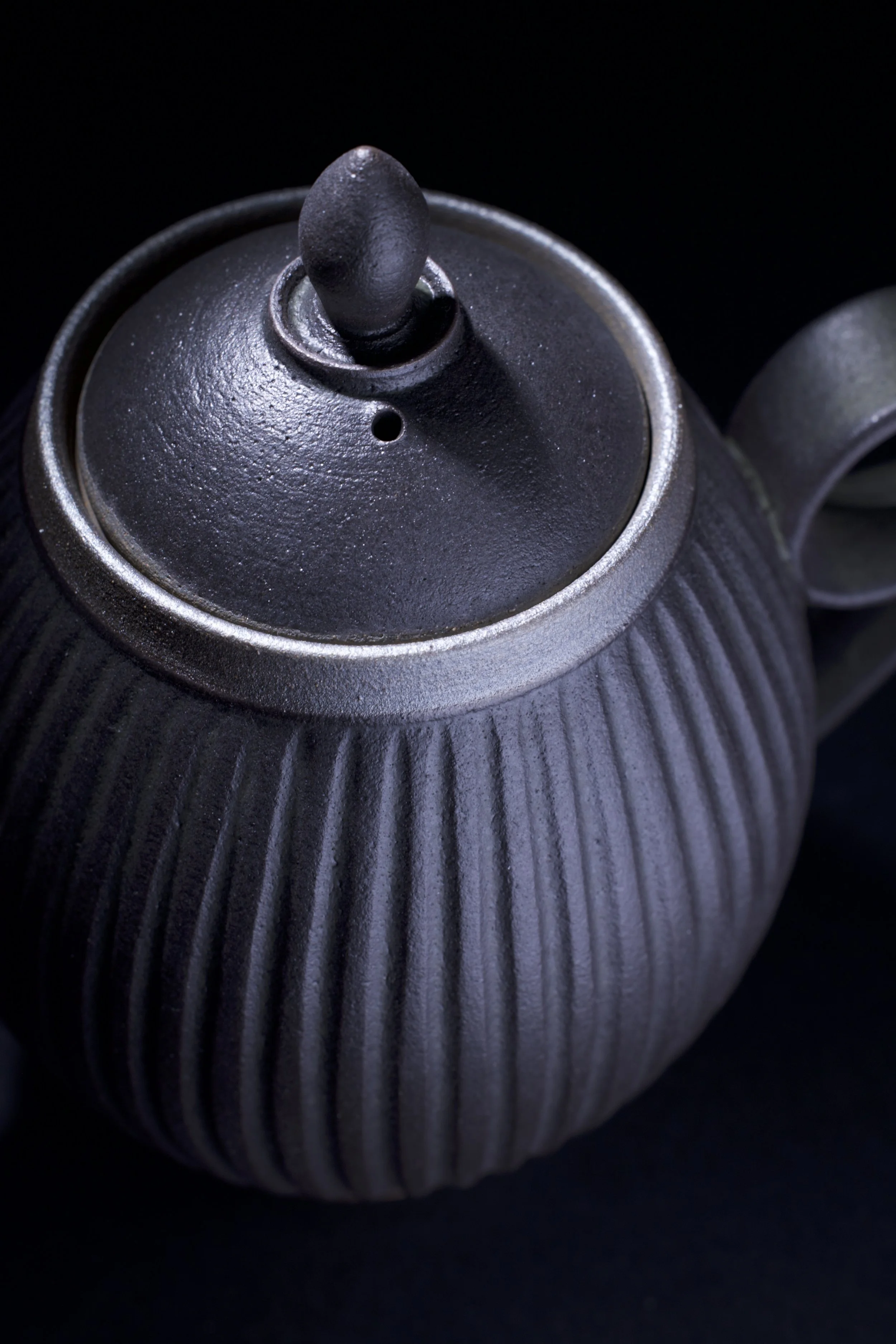 Close-up of a dark gray matte ceramic teapot with a textured ribbed surface, set against a black background.