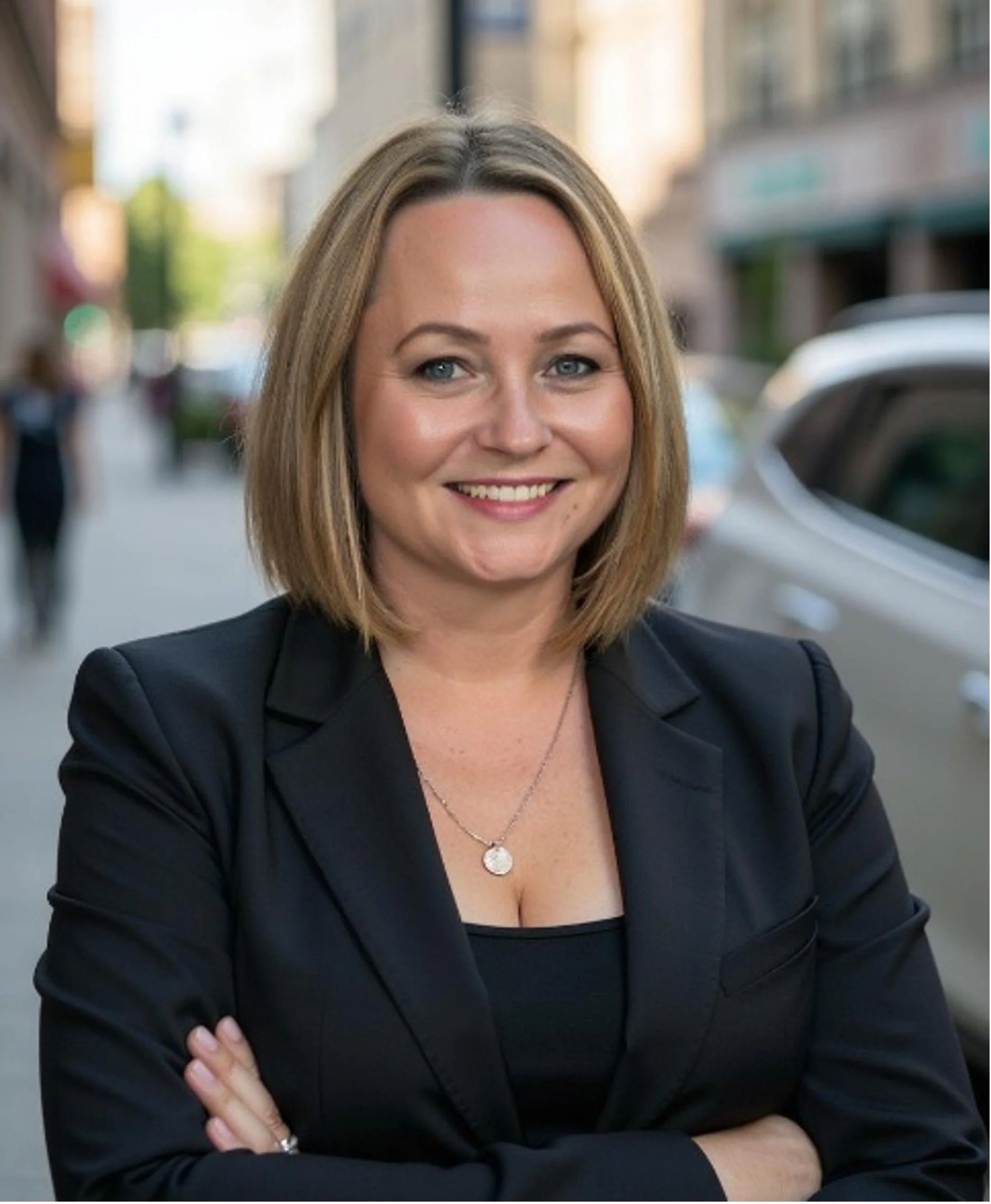 Pam Black, in black blazer smiling with arms crossed on a city street