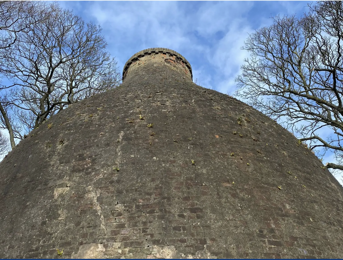 Perspective view of a large, round stone structure with a turret, surrounded by leafless trees under a blue sky.