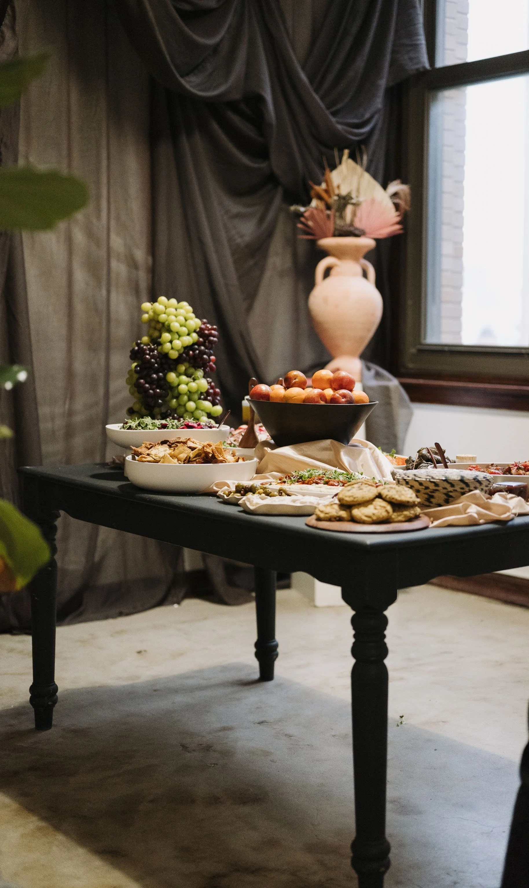 Black table of a mezze spread with grape tower, wooden bowl filled with oranges, with black draping and large white vase with dry flowers