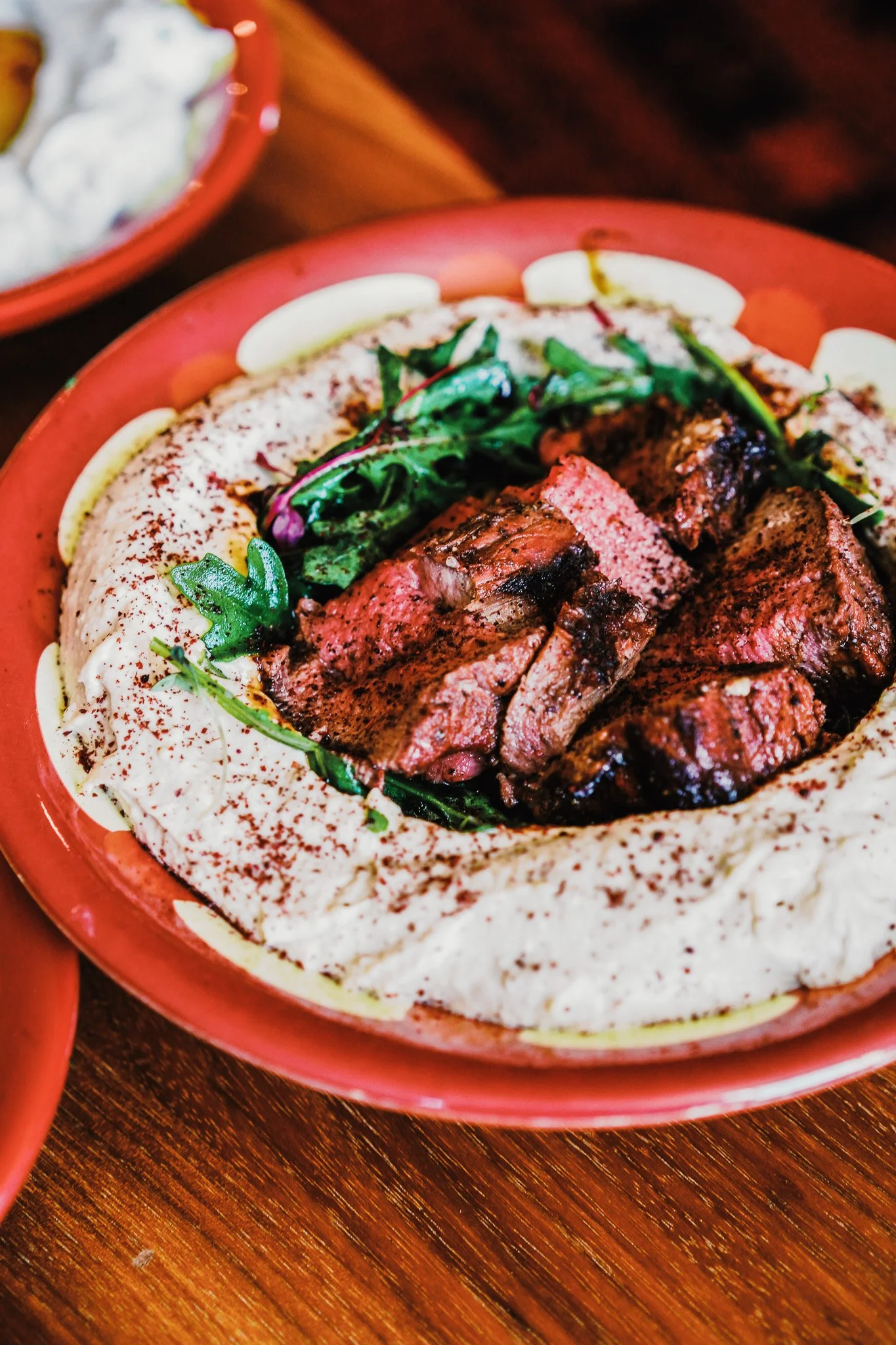 bowl of hummus with a sprinkle of sumac with chunks of beef tenderloin and arugula leaves