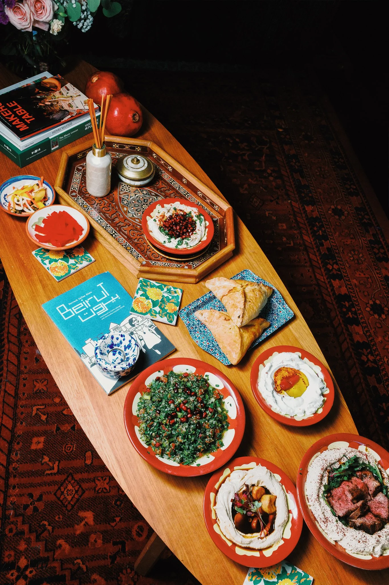 Mezze table with traditional bowls filled with hummus, labneh, tabbouleh, and pickles. Around it are traditional and modern tiles, trays, and books. 