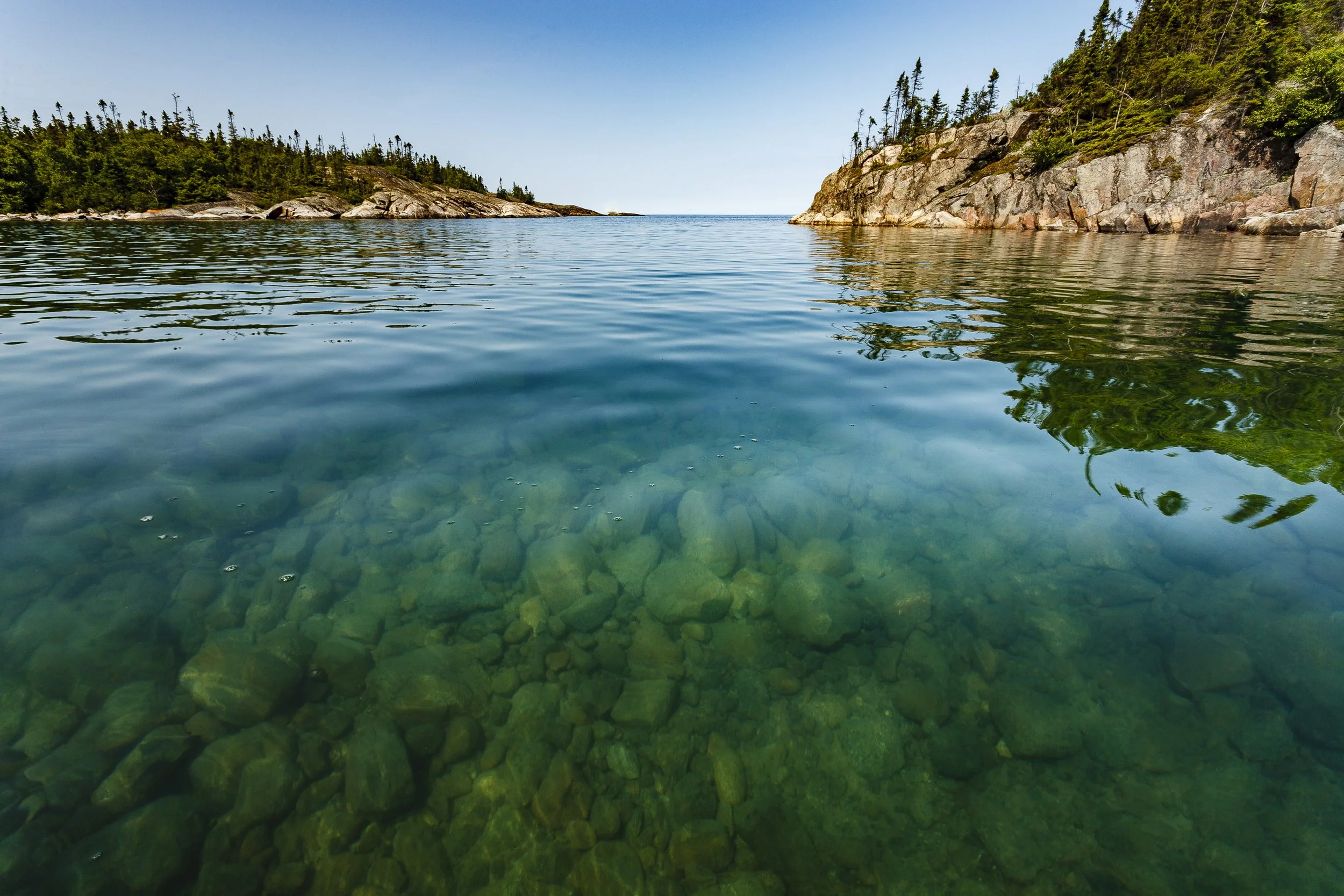 Clear water with visible rocks at the bottom, surrounded by forested rocky cliffs under a bright sky.