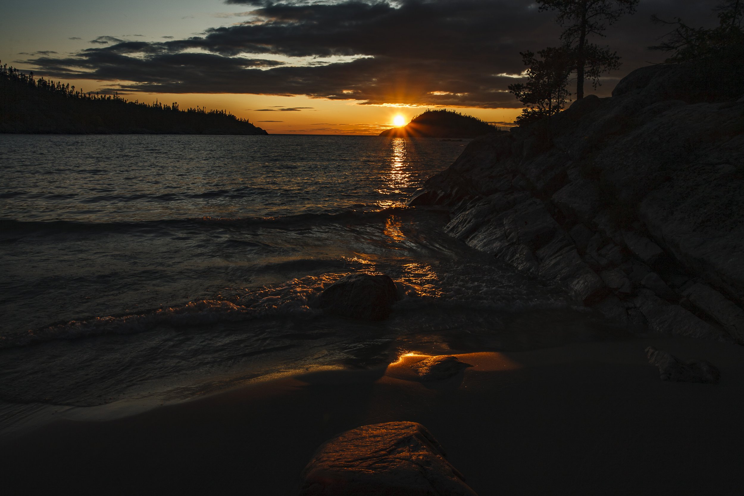 Sunset over a lake with dark clouds, reflecting orange and yellow hues on the water, with rocky shoreline and silhouetted trees in the background.