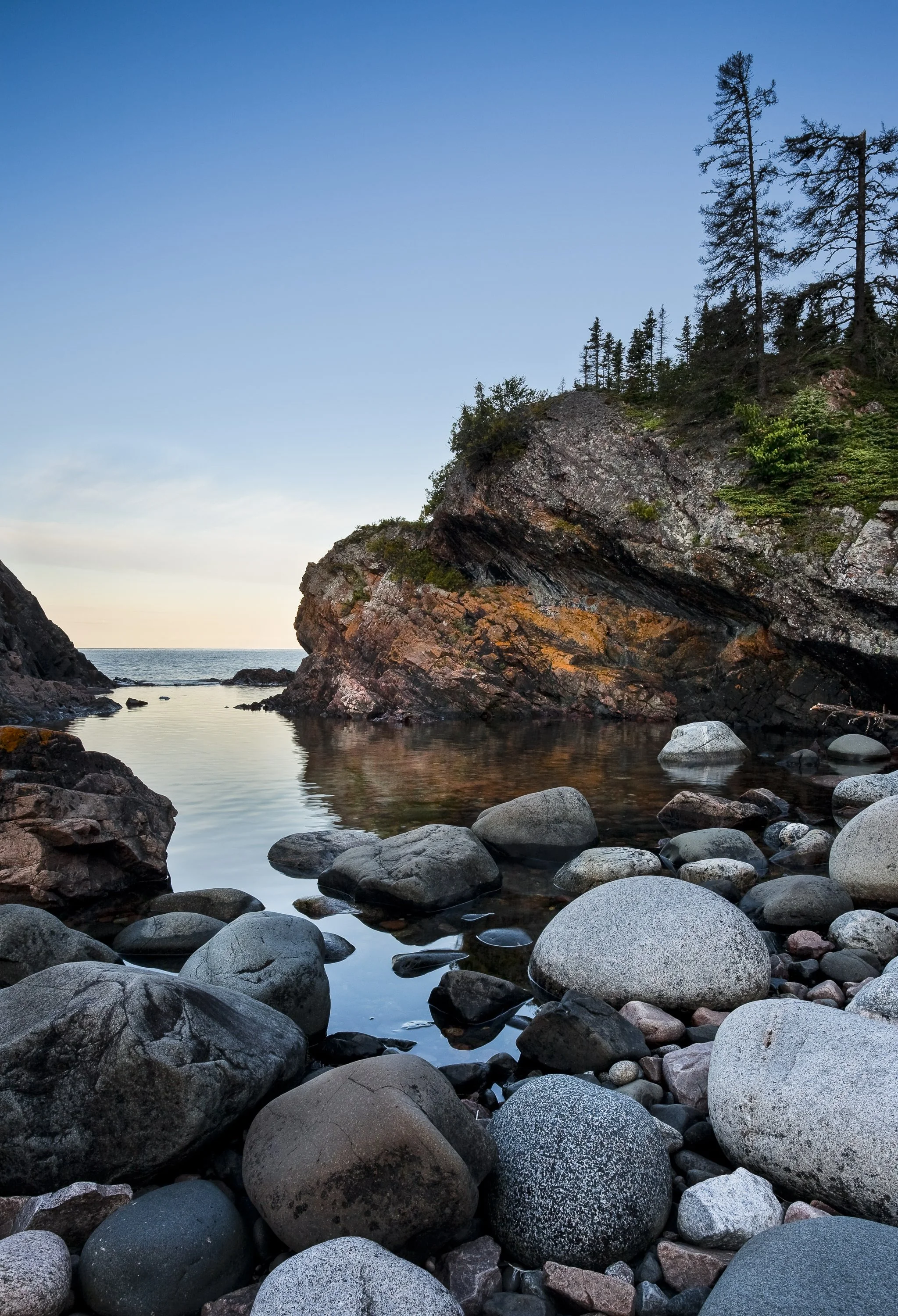 A rocky coastal scene with large smooth stones in the foreground, calm water reflecting the rocks, steep cliffs with sparse trees on top to the right, and an ocean horizon in the background under a clear blue sky.