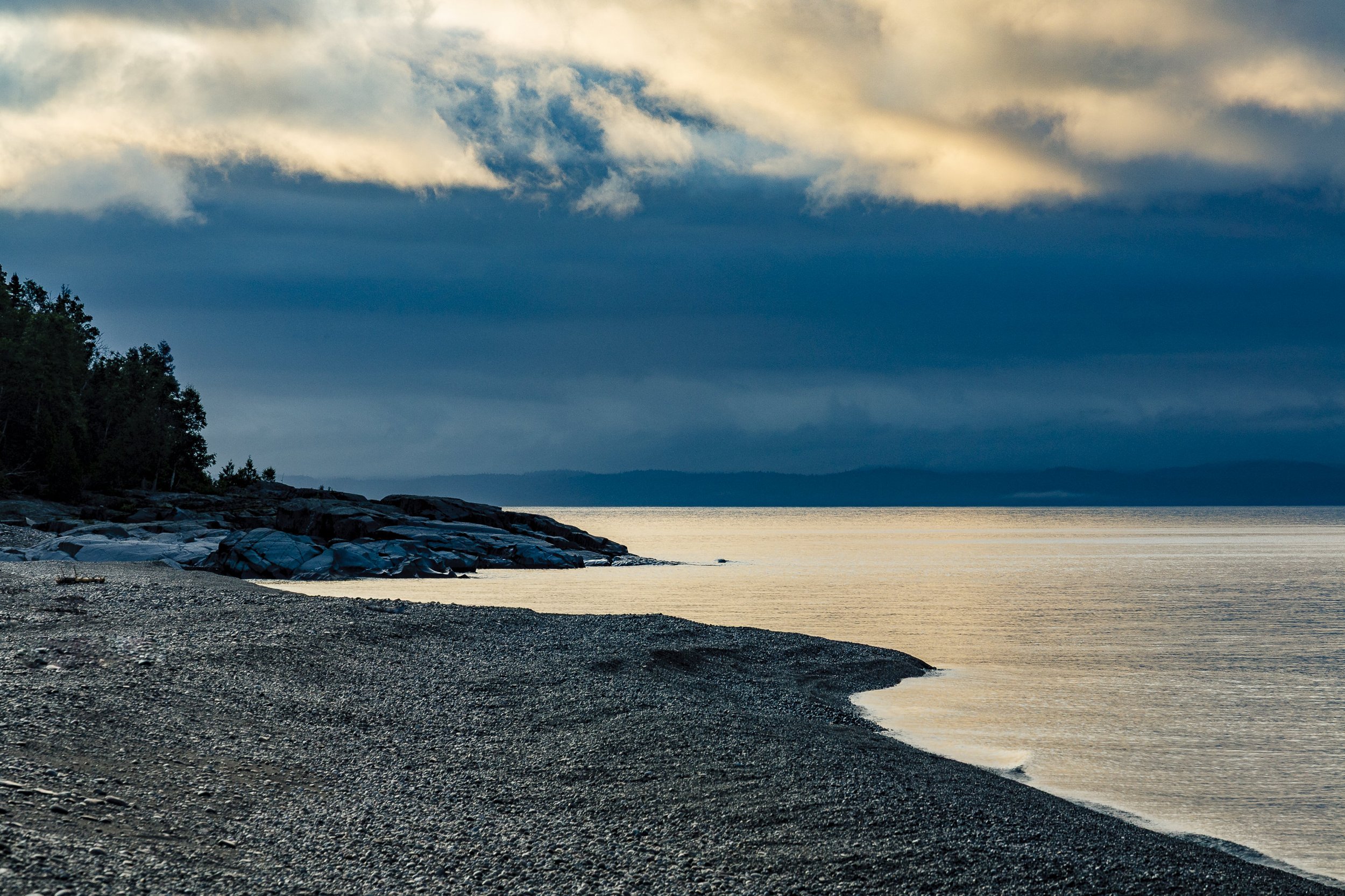 A rocky beach at dusk with a forested shoreline on the left, dark clouds overhead, and the calm ocean reflecting the muted sky.
