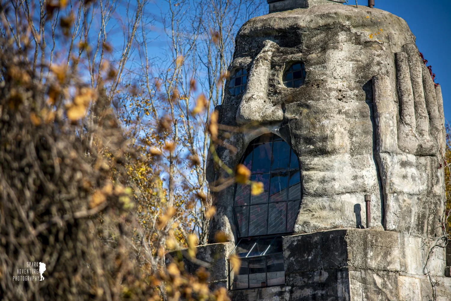 A stone monument in the shape of a person's face, with a large open mouth, round eyes, and a nose, surrounded by trees with autumn leaves and a clear blue sky.