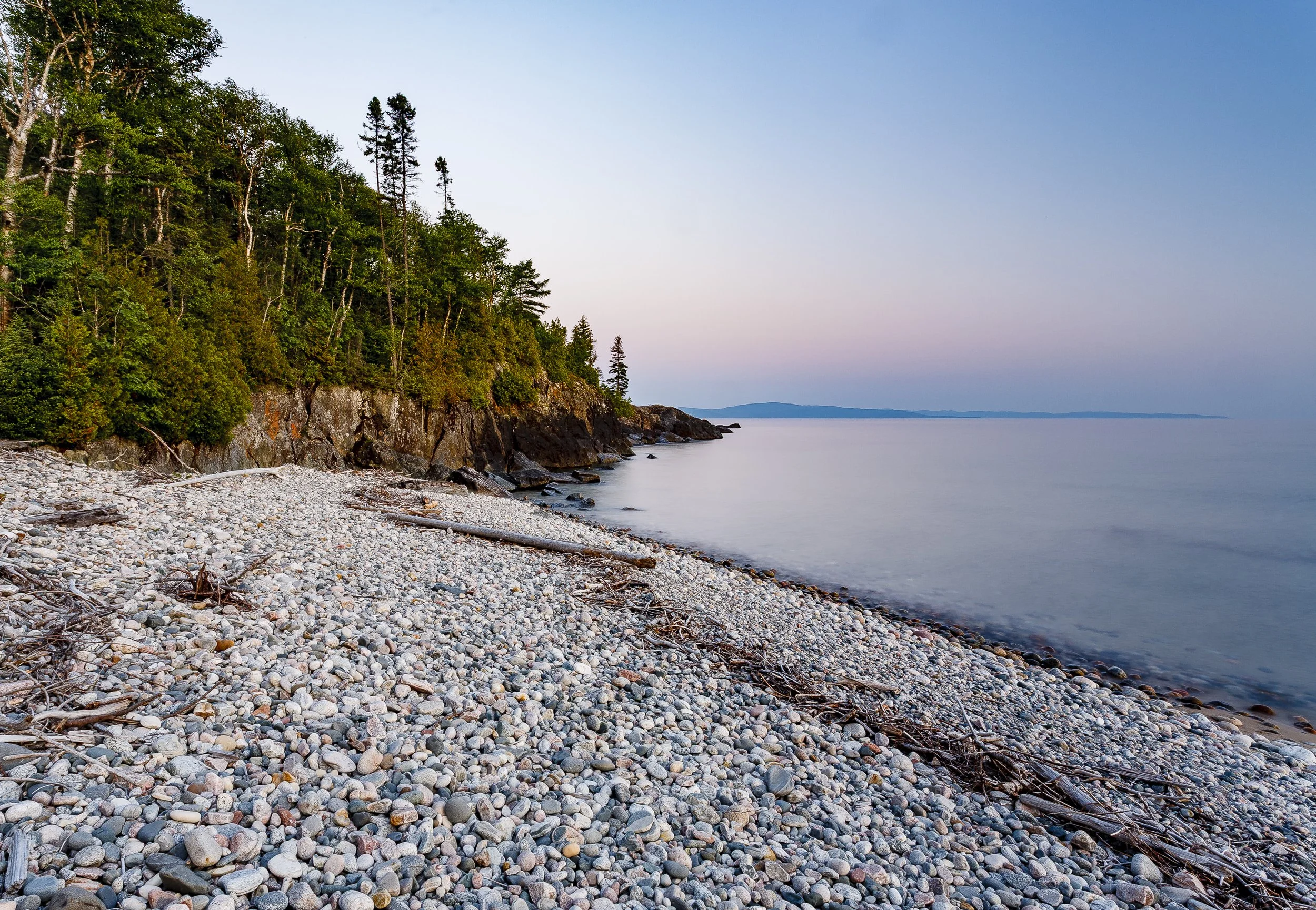 A rocky beach with a wooded cliffside on the left and calm water extending to the horizon with a faint landmass in the distance during dusk.