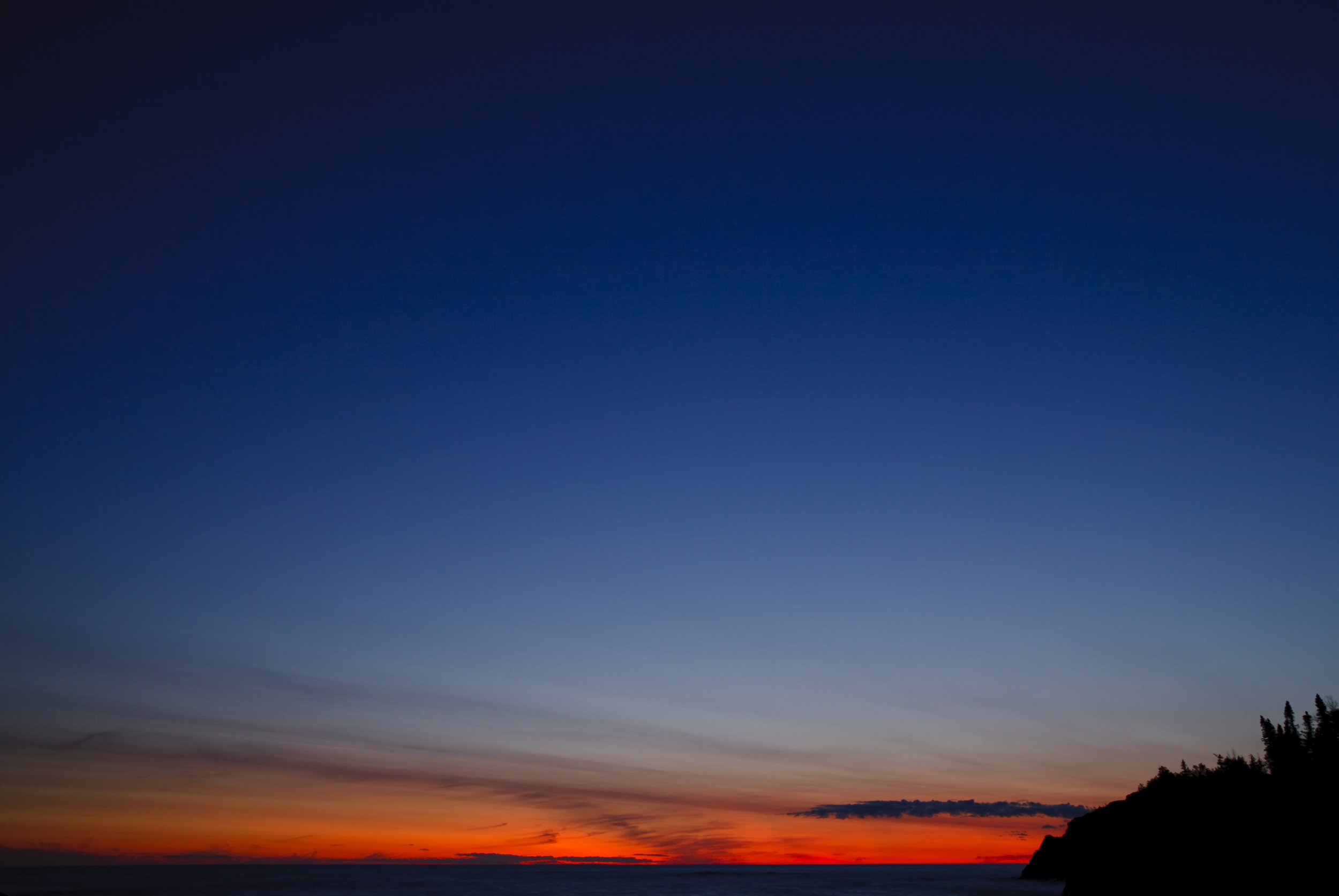 Sunset over the ocean with a silhouette of a cliff on the right and a mostly clear sky transitioning from deep blue at the top to warm orange near the horizon.