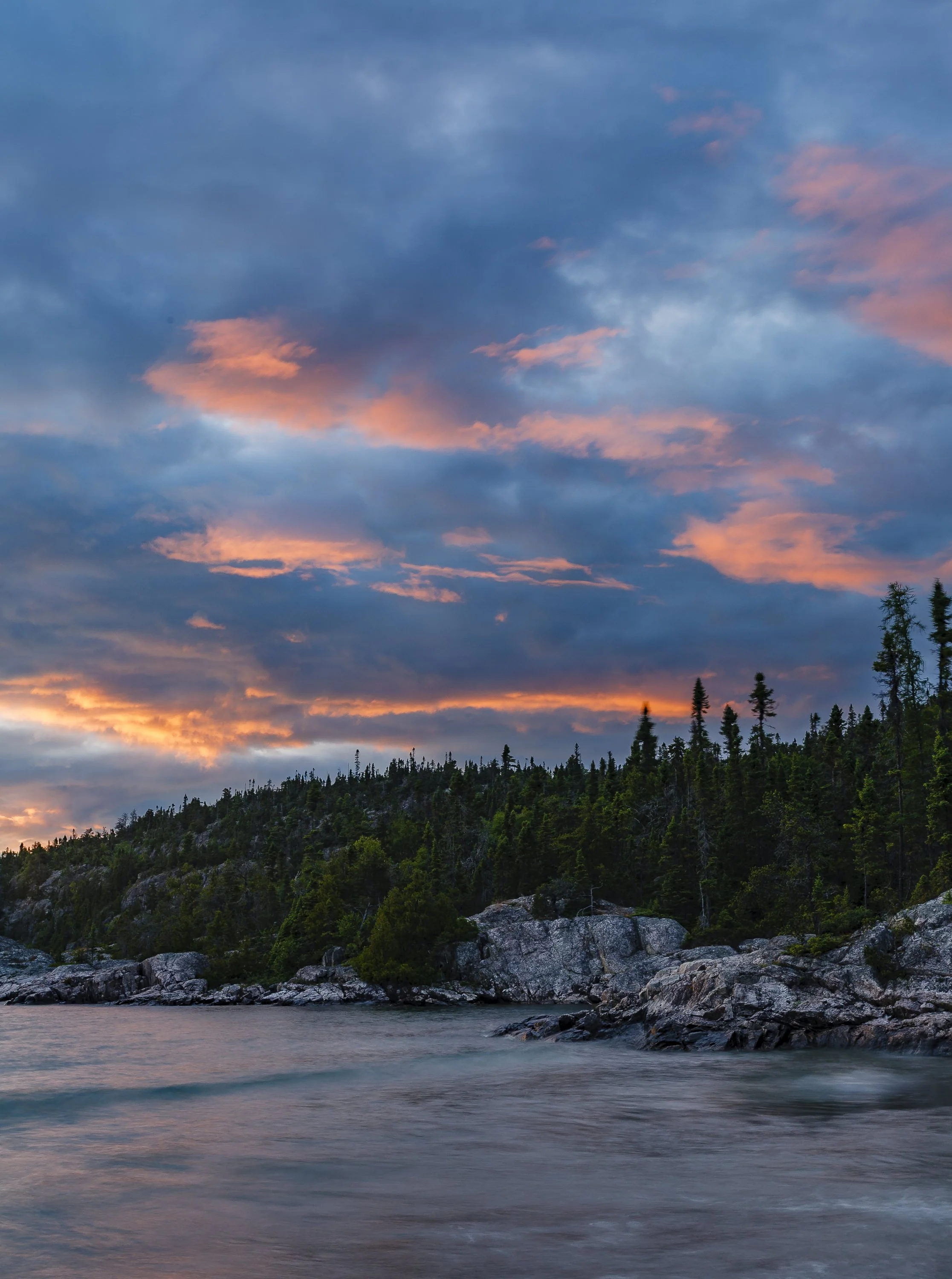 A landscape at sunset with a cloudy sky, a forested hillside, and a body of water in the foreground.