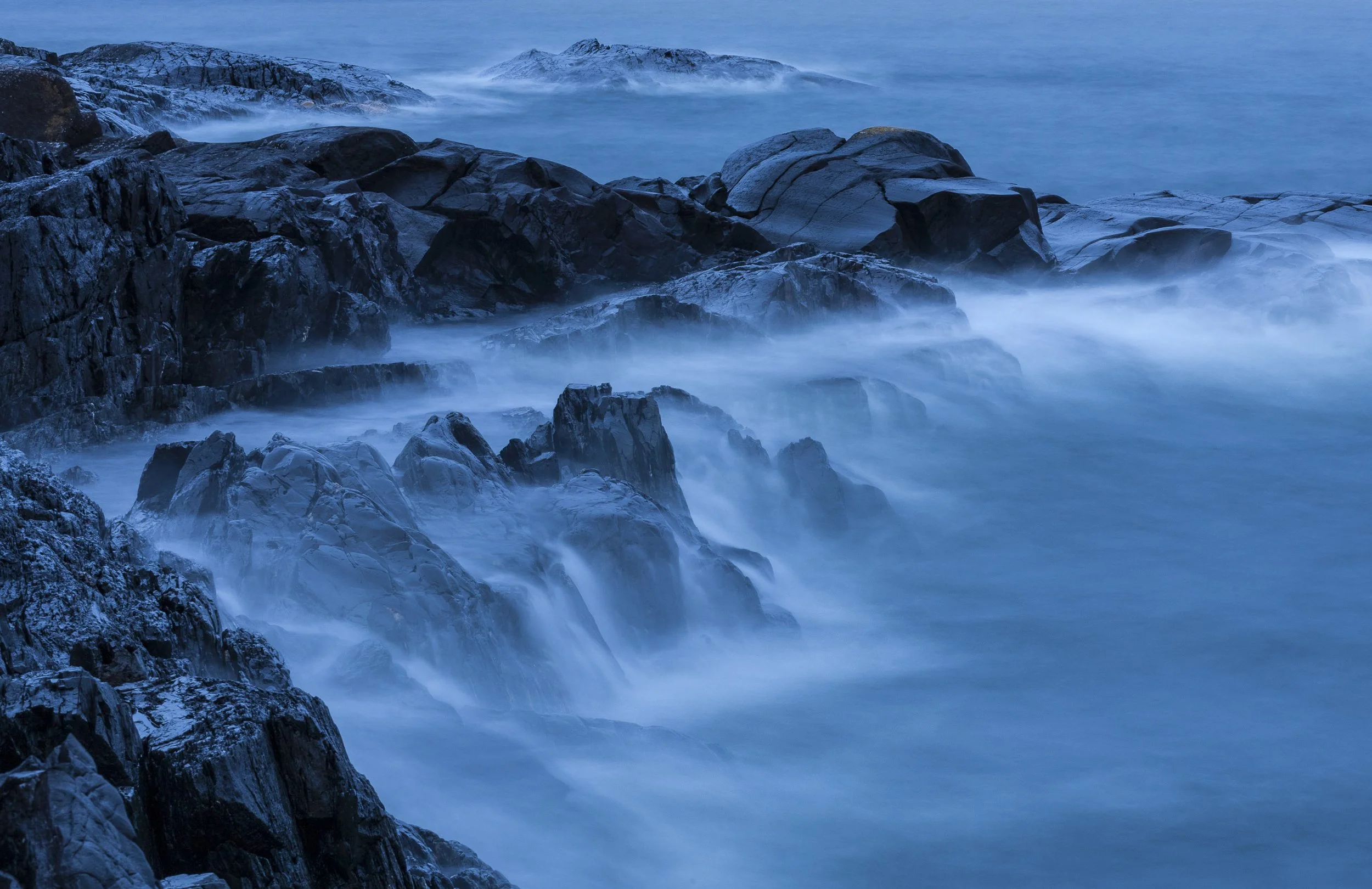 Long exposure photograph of rugged rocky coastline with waves crashing against the rocks, misty water flowing around the rocks, and distant snow-capped mountains in the background under a blue sky.