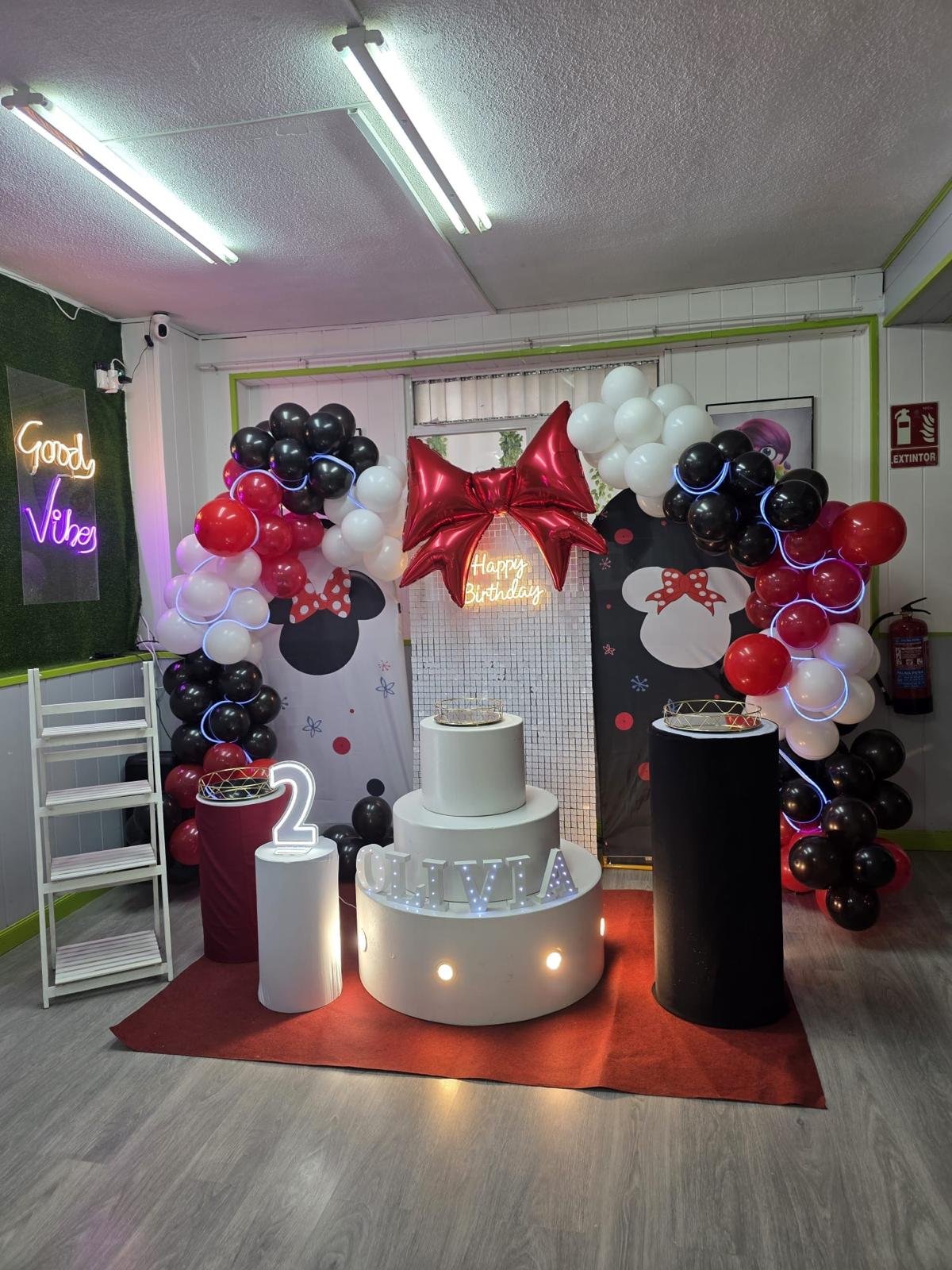 Decoración de cumpleaños con globos en colores negro, blanco y rojo, con un arco de globos, una luz LED que dice 'Happy Birthday', una dona en una mesa, y letras que dicen 'Olivia' en uno de los pedestales.