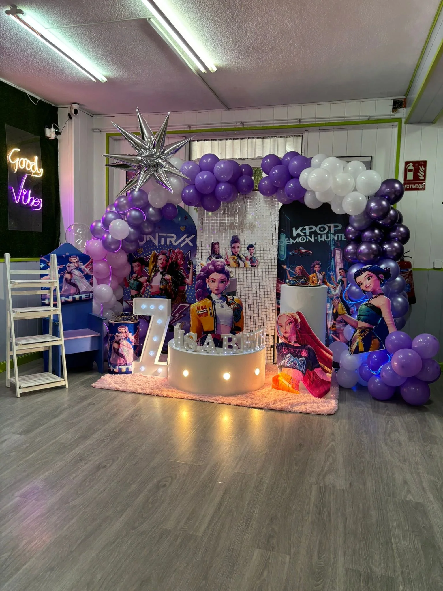 Decoración para una fiesta infantil con globos morados, blancos y plateados. Incluye un arcor de estrellas plateadas, personajes de dibujos animados y letras luminosas que dicen '7 SAB'.