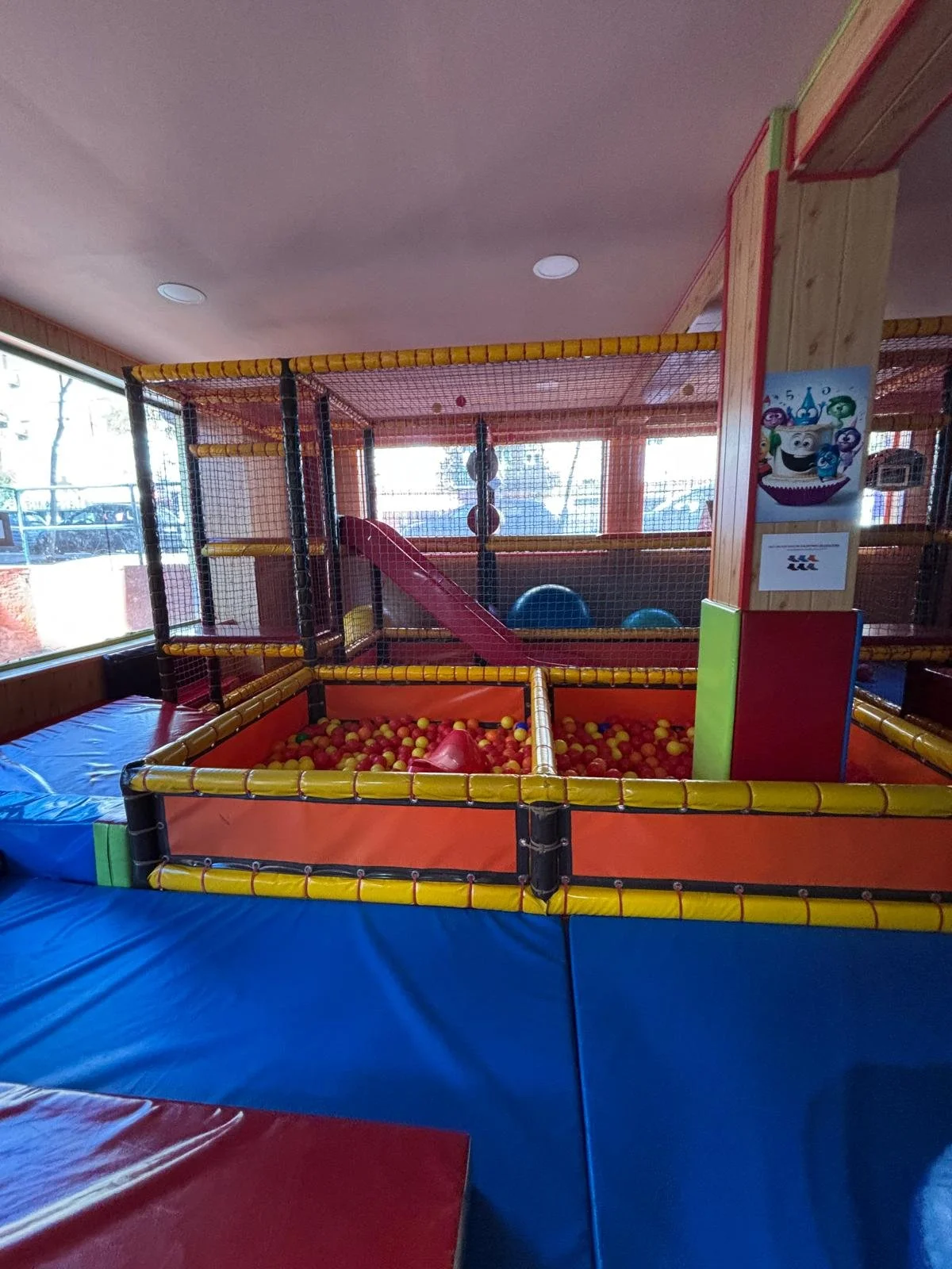 Área de juego infantil con un castillo de pelotas con redes de protección y resbaladilla, en un espacio cerrado con paredes de vidrio.