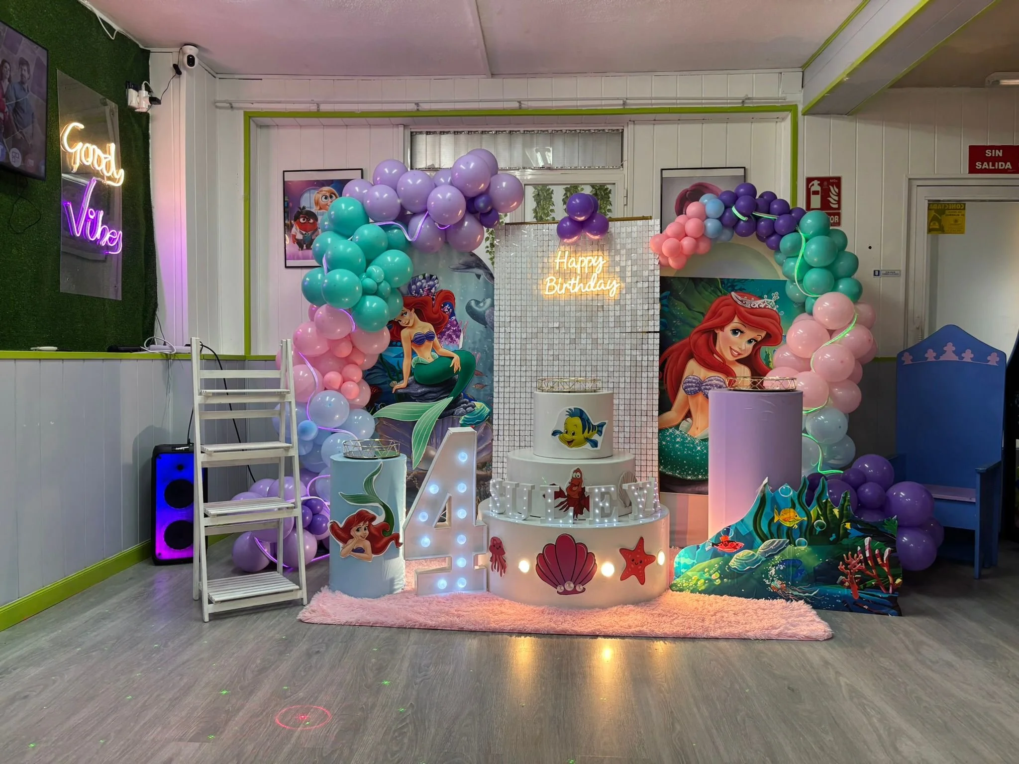 Decoración de cumpleaños con temática de sirenas, globos de colores pastel, carteles y figuras de Ariel, y un cartel que dice 'Feliz Cumpleaños'.