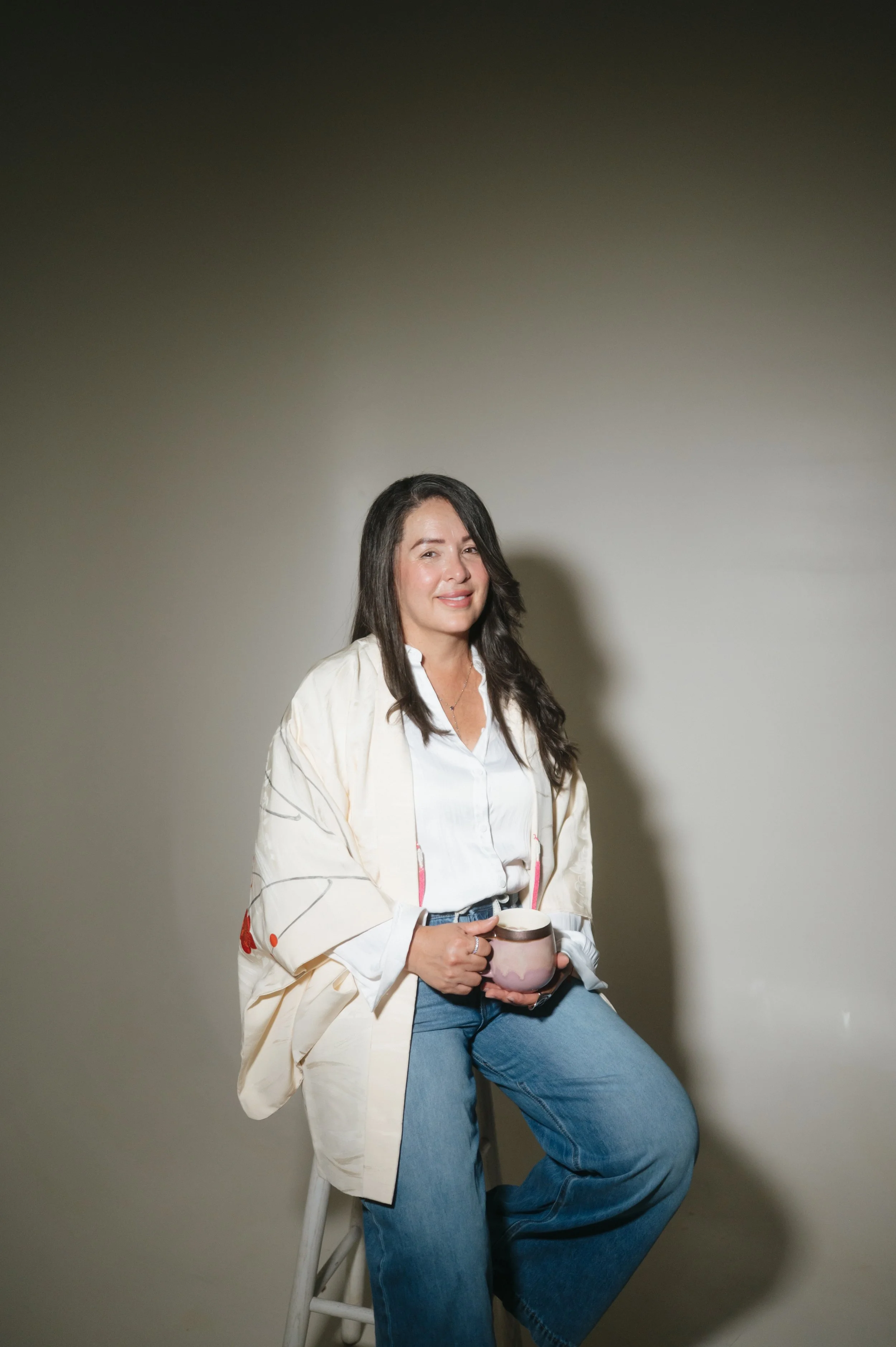 A woman with long dark hair sitting on a white stool, smiling, holding a mug, wearing a white shirt and blue jeans, against a plain gray background.