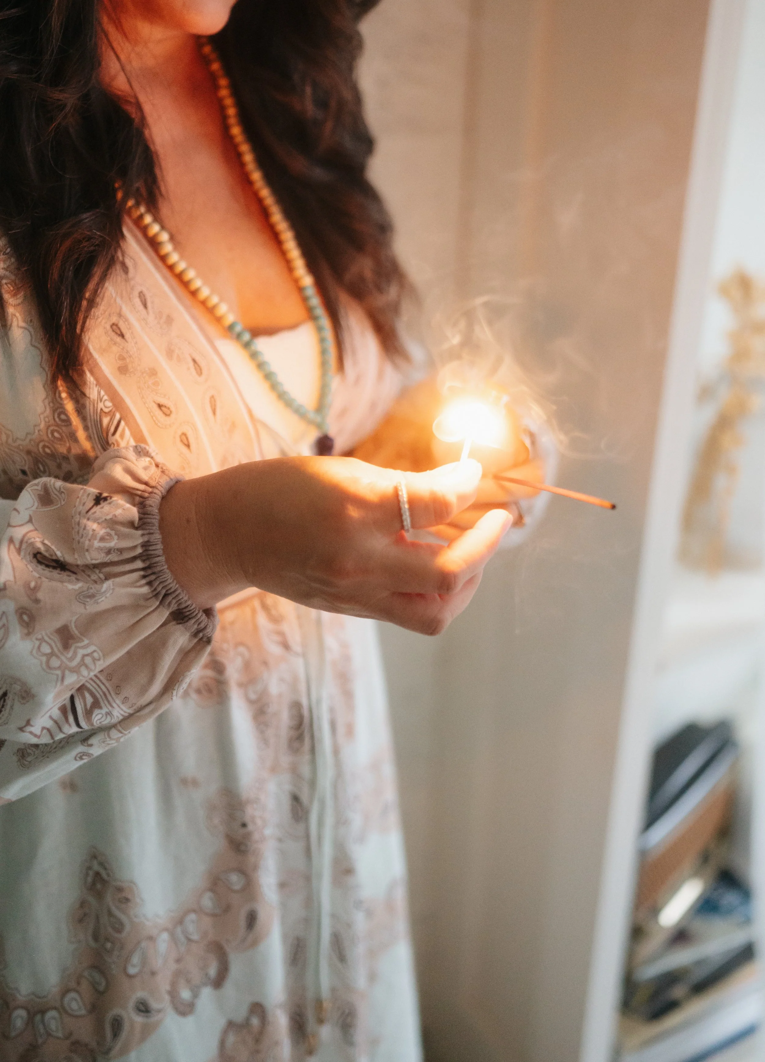 A woman holding a lit matchstick, with visible smoke, near a candle or small light source, wearing a patterned blouse and beaded necklace.