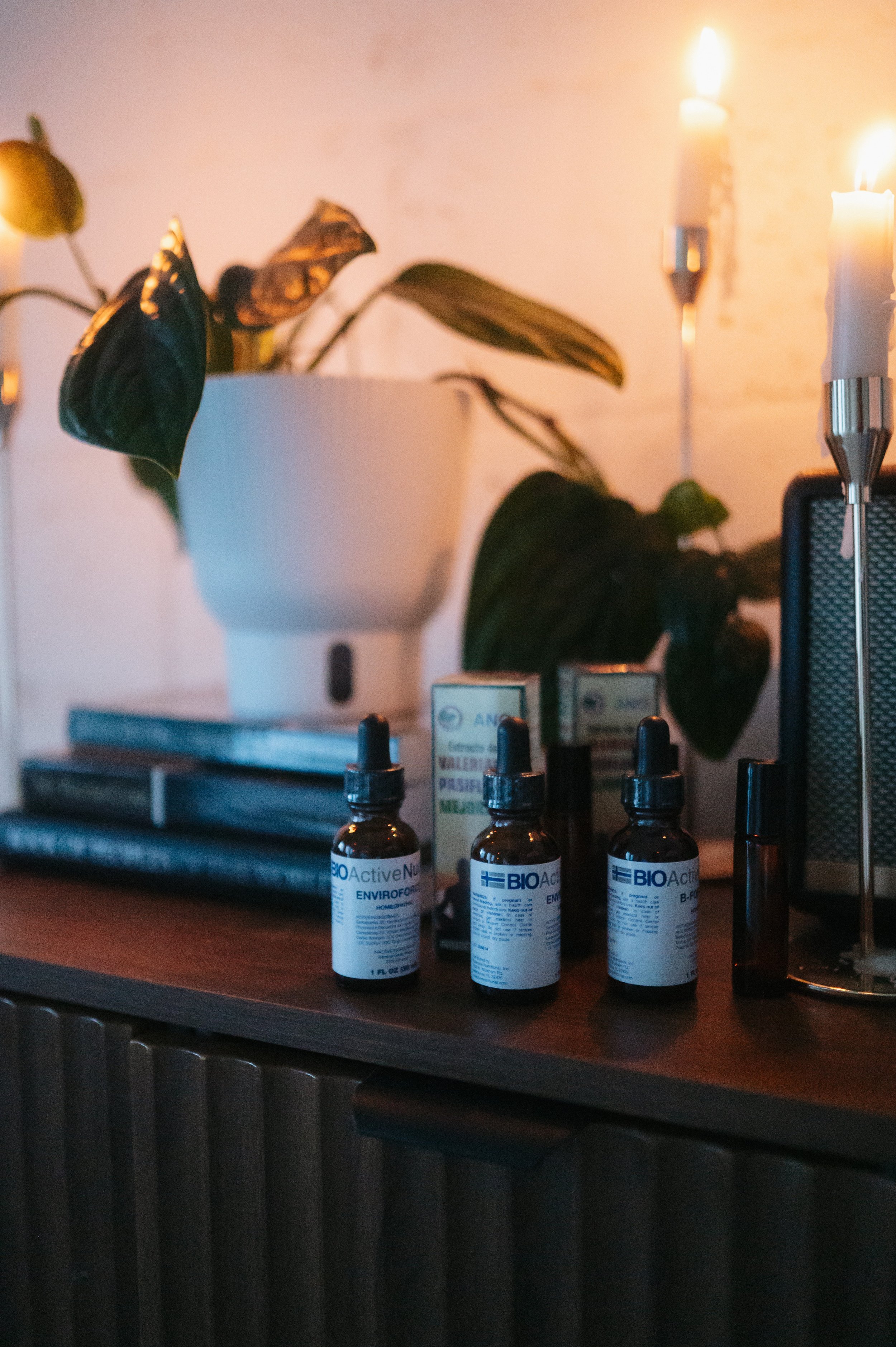 A wooden table with three small brown bottles labeled BIO Active N, a modern set of candles with bright flames, a potted plant with green leaves, and a black speaker, with some blurred objects in the background.