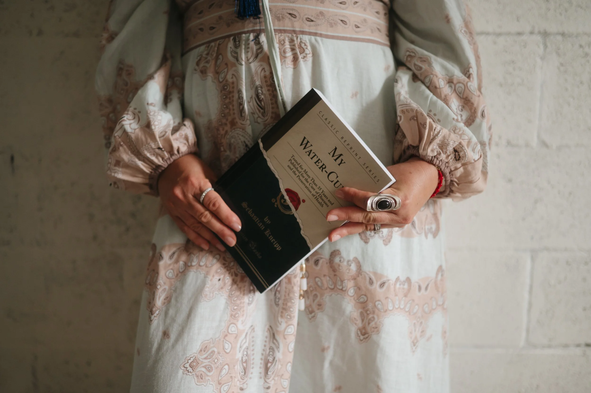 Person wearing a pink and white patterned dress holding a book titled "My Water Cure" in front of a white brick wall.