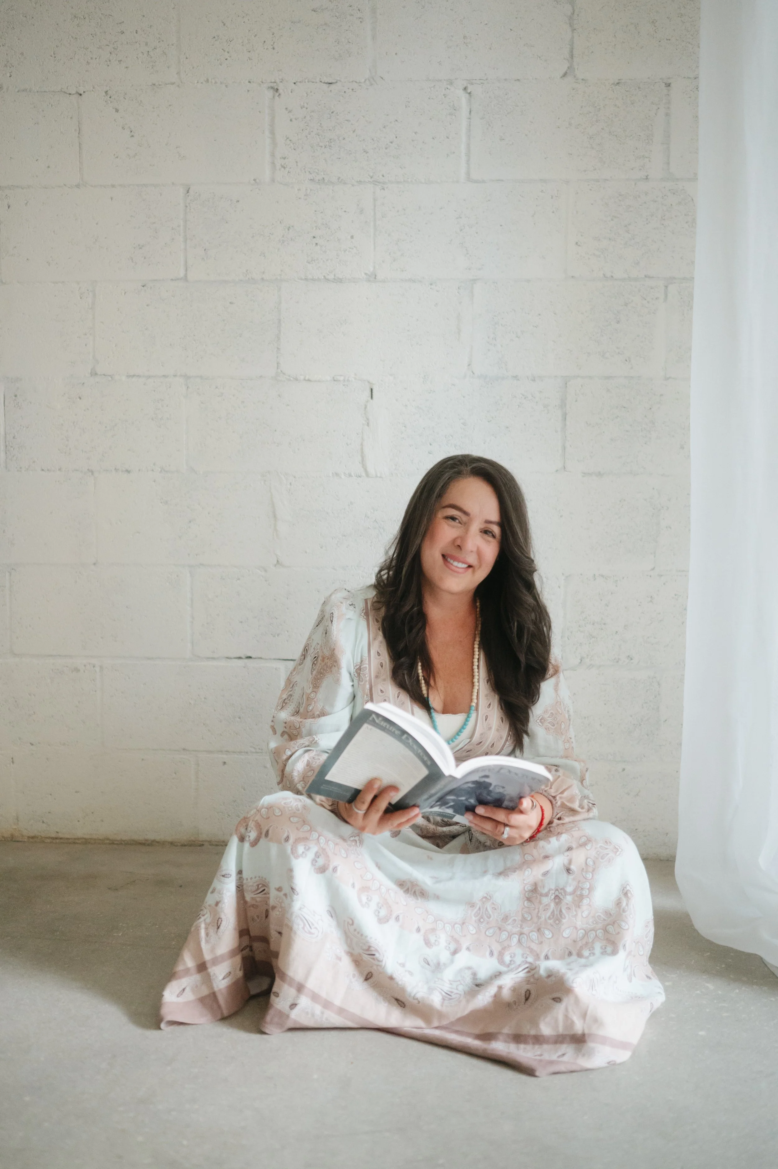 A woman with long dark hair wearing traditional white and pink embroidered dress, sitting on the floor, holding and reading a book, in front of a white brick wall, next to a white curtain.