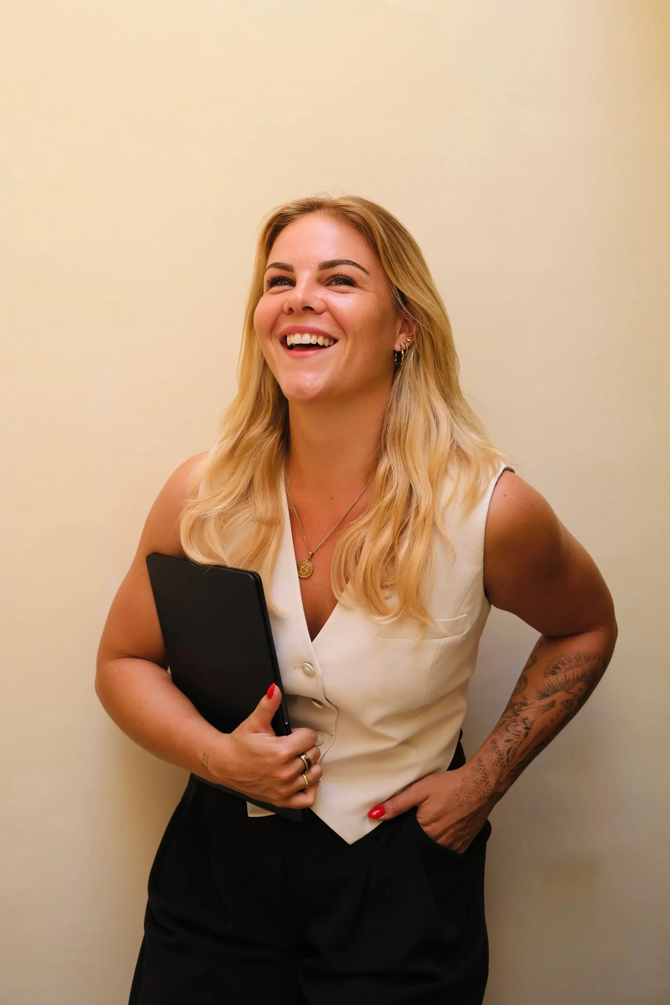 co-founder paula, shoulder length blonde hair, white top, black pants, holding laptop, beige background, smiling