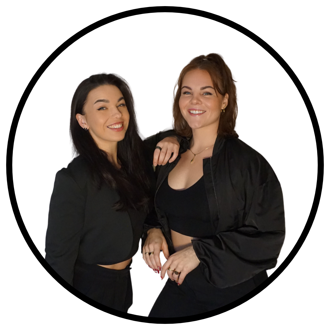 Two smiling sisters with dark and light brown hair, wearing black outfits and jackets, standing close together against a white background